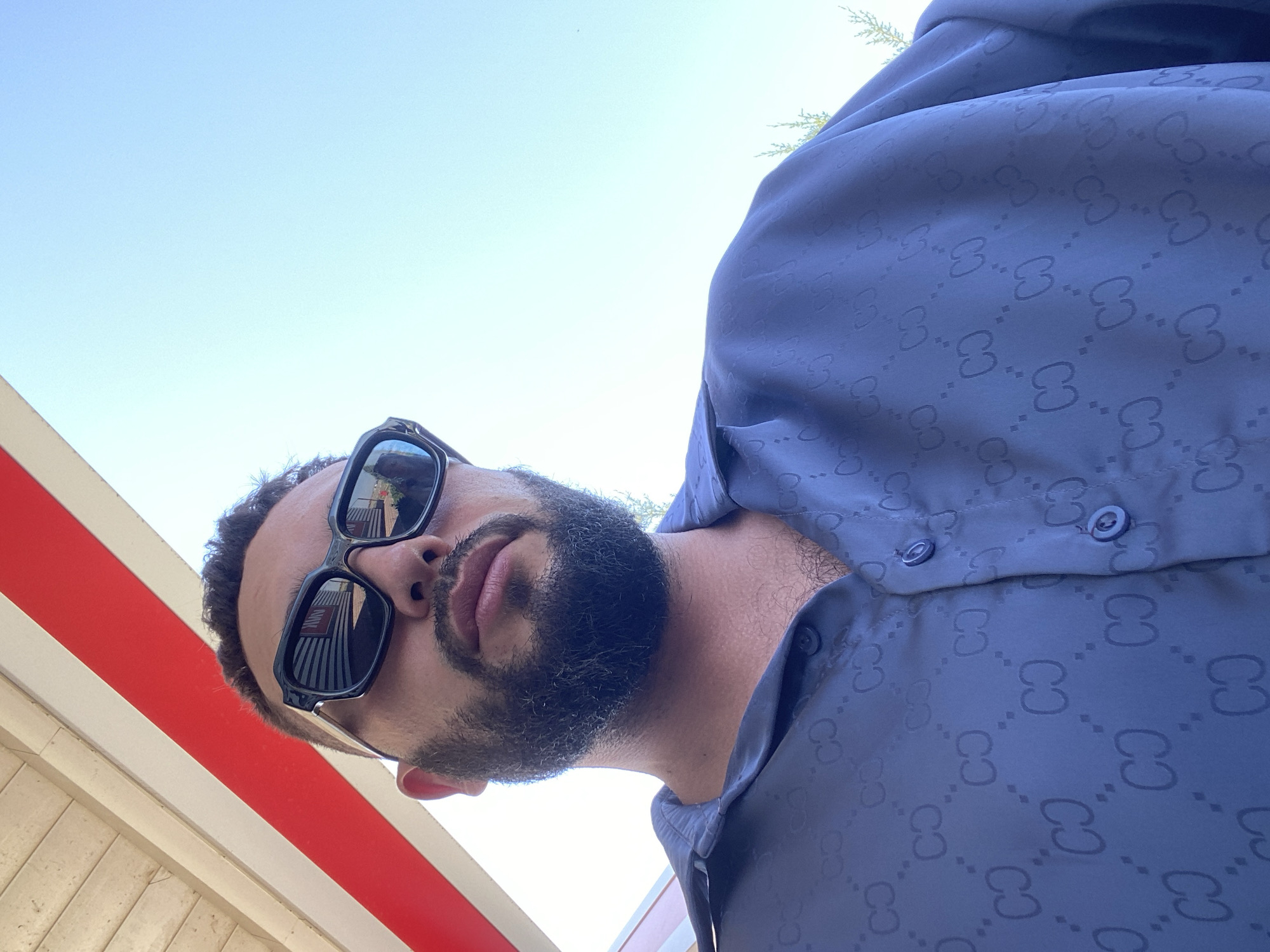 A man wearing sunglasses and a blue shirt with a repeating pattern is pictured. He has a beard and is looking away from the camera. The background consists of a blue sky and a red and white striped structure. The location is not identifiable within the city of Korkmaskala, Russia.