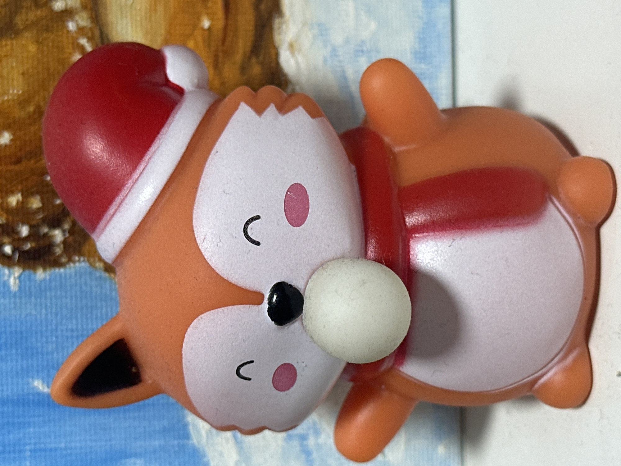 A close-up, eye-level shot shows a small, orange fox figurine wearing a red and white Santa hat and a red and white striped scarf. The figurine has a white face with closed eyes and a black nose, and it is holding a white, spherical object in its mouth. The figurine is positioned in front of a blurred background that appears to be a winter scene with a blue sky and a snow-covered landscape. There is no information about the location in Nevinnomyssk, Russia.