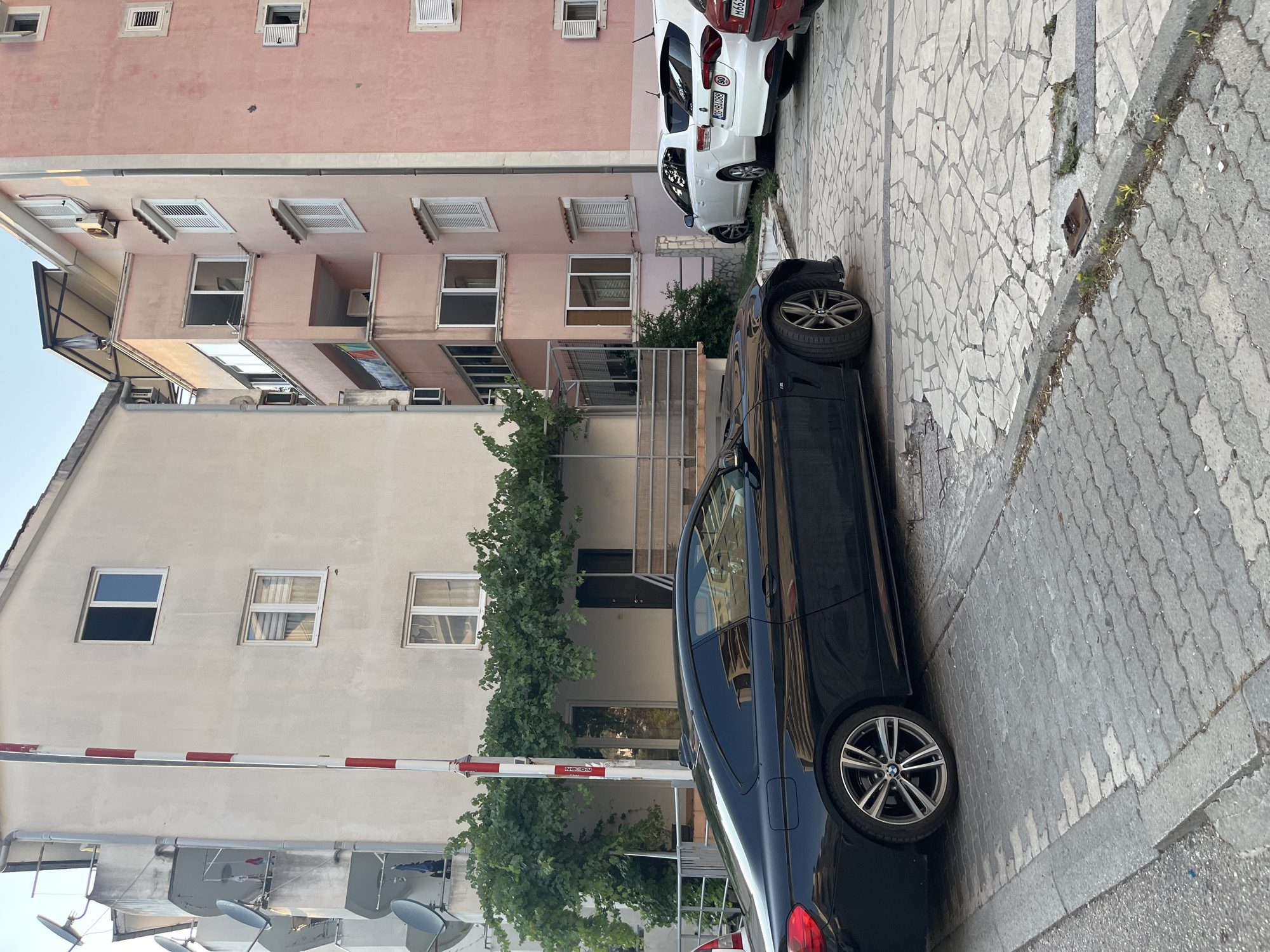 This image depicts a street scene in Budva, Montenegro. A black BMW is parked along a narrow cobblestone street. Adjacent to the street is a light-colored building with several windows and a red and white striped barrier. Further back, a pink building with multiple windows and air conditioning units is visible. A white car is parked in front of the pink building. The scene suggests it is daytime, likely late afternoon, with clear weather. The presence of parked cars and the architecture point to an urban residential area. There are no people visible in the image. Some faint text is visible on the barrier, but it is illegible.