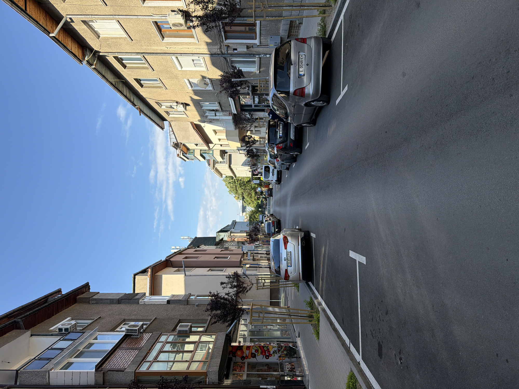 The image shows a street scene in Blagoevgrad, Bulgaria. The view is down a residential street with buildings on either side. Several cars are parked along the sides of the street, lined up in designated parking spots marked by white lines. The street is paved with asphalt. The sky is blue with a few scattered clouds. There are small trees planted along the sidewalk, and the buildings are of varying heights and architectural styles.