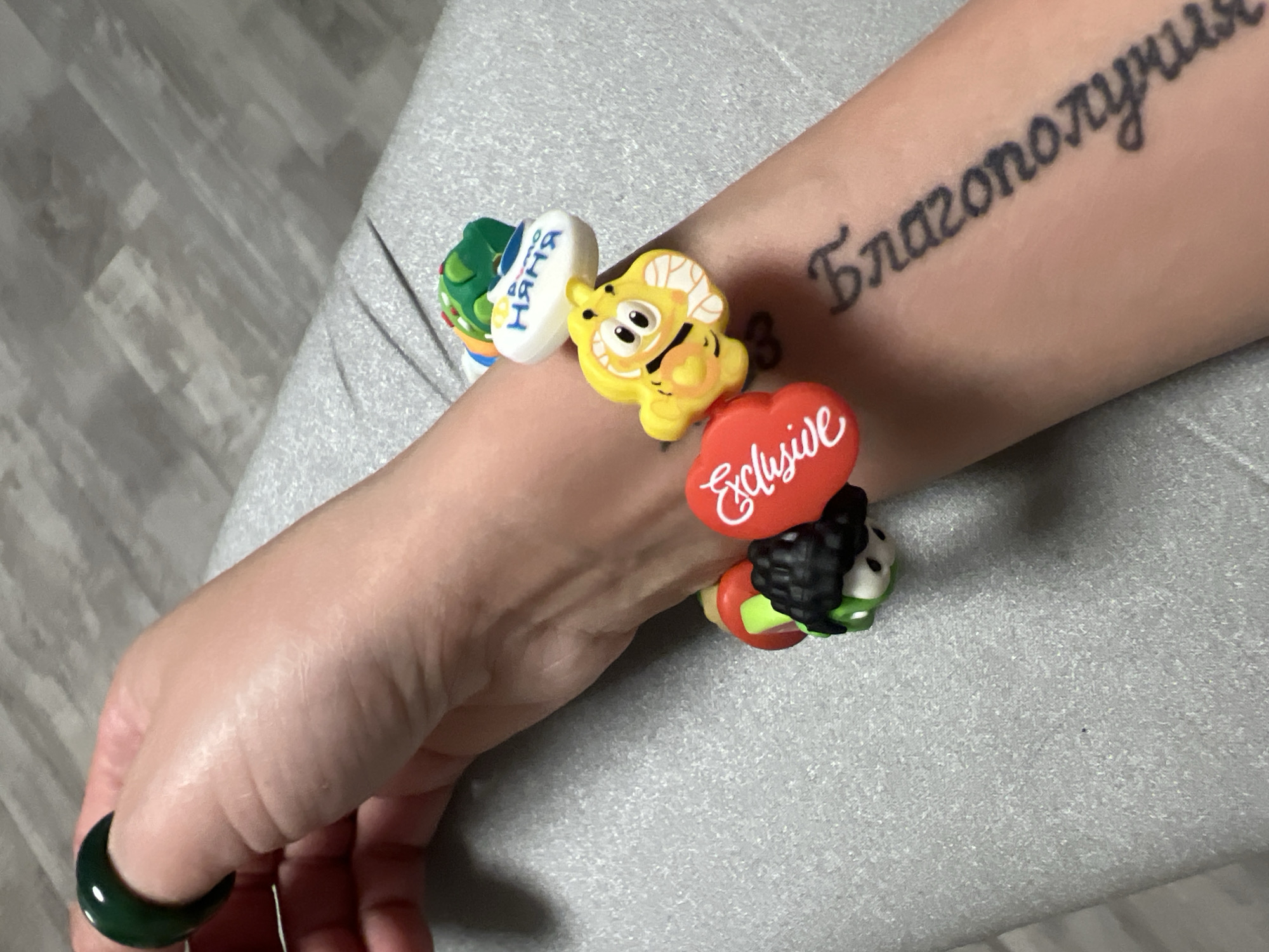 A close-up shot displays a fair-skinned arm adorned with a beaded bracelet featuring colorful, whimsical charms, positioned against a soft gray backdrop. The tattoo reads "Благополучия".
The finger on the left hand has a dark green ring.
The scene's location is unknown.