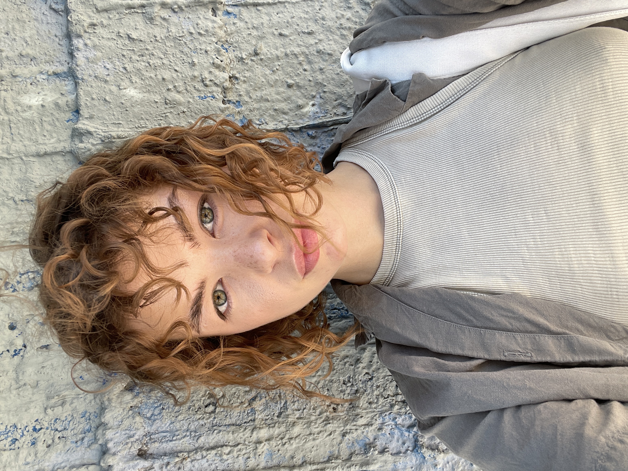 A young woman with curly red hair, fair skin, and green eyes is lying on a textured gray and blue painted surface, possibly a wall or ground. She is wearing a light gray striped shirt and a dark gray hooded sweatshirt. The woman's face is tilted towards the viewer. The location cannot be determined from the image.