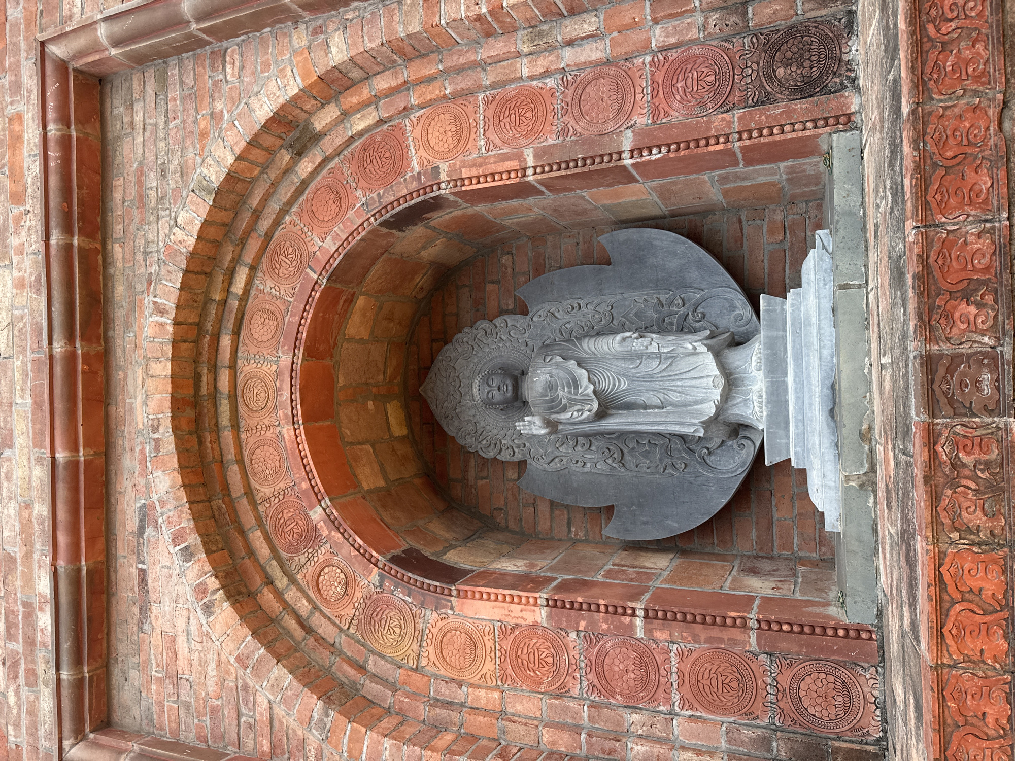 The image shows a Buddha statue standing in an alcove within a brick structure. The alcove is arched and decorated with circular motifs carved into the bricks. The Buddha statue is made of grey stone and is depicted standing with its right hand raised. The base of the statue is a tiered platform. The surrounding brickwork is aged and shows variations in color. The scene is outdoors, and the lighting suggests daytime. The location is identified as Tây Hoa Lư Ward, Vietnam.