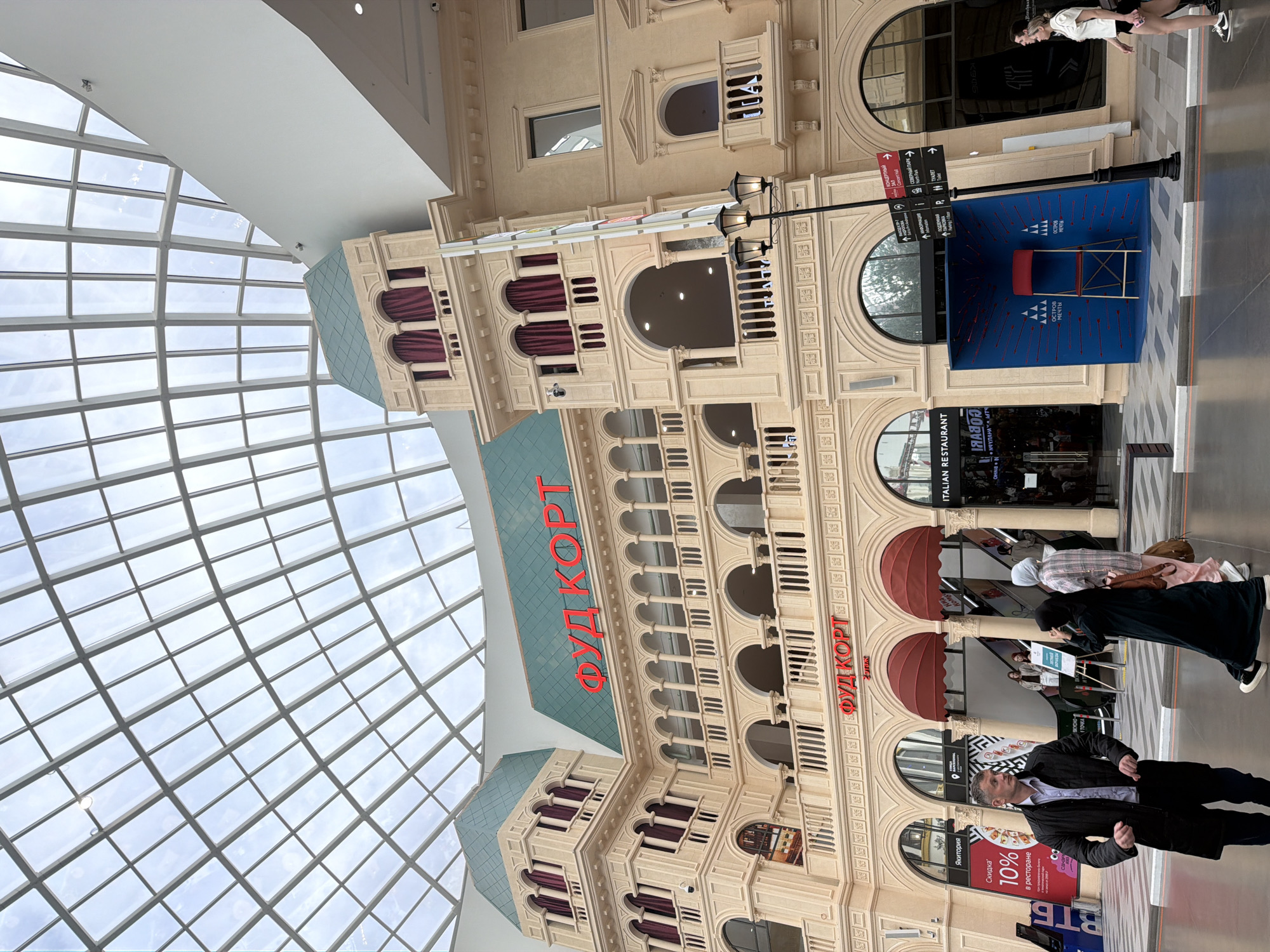 This is a medium shot, rotated, of a brightly lit indoor space, likely a shopping mall, with a glass ceiling. The architecture features a faux classical facade with ornate detailing, arches, columns, and decorative moldings in a light beige color. Red velvet drapery is visible behind some of the arched windows. A large sign with red Cyrillic lettering that reads "ФУД КОРТ" (Food Court) is prominent on the building.
In the foreground, several people are visible, some walking and some standing. One man in a dark jacket and lighter shirt is looking towards the camera. A woman in a white top and black shorts is captured in motion, appearing to run or walk briskly. Other individuals are seen further back, some with head coverings.
A blue display box with red arrows pointing inwards and a red chair in the center is positioned near a lamppost with three lights. Signs with Cyrillic text are mounted on the lamppost, indicating directions to various locations and amenities like a concert hall, restrooms, and parking. One sign prominently features the words "ОСТРОВ МЕЧТЫ" (Island of Dreams).
A retail advertisement for "Якитория" offering a "10% discount in the restaurant" is visible on a pink banner. The overall atmosphere appears to be one of a busy commercial environment. The time of day is indeterminate due to the indoor setting, but the lighting is bright and even.