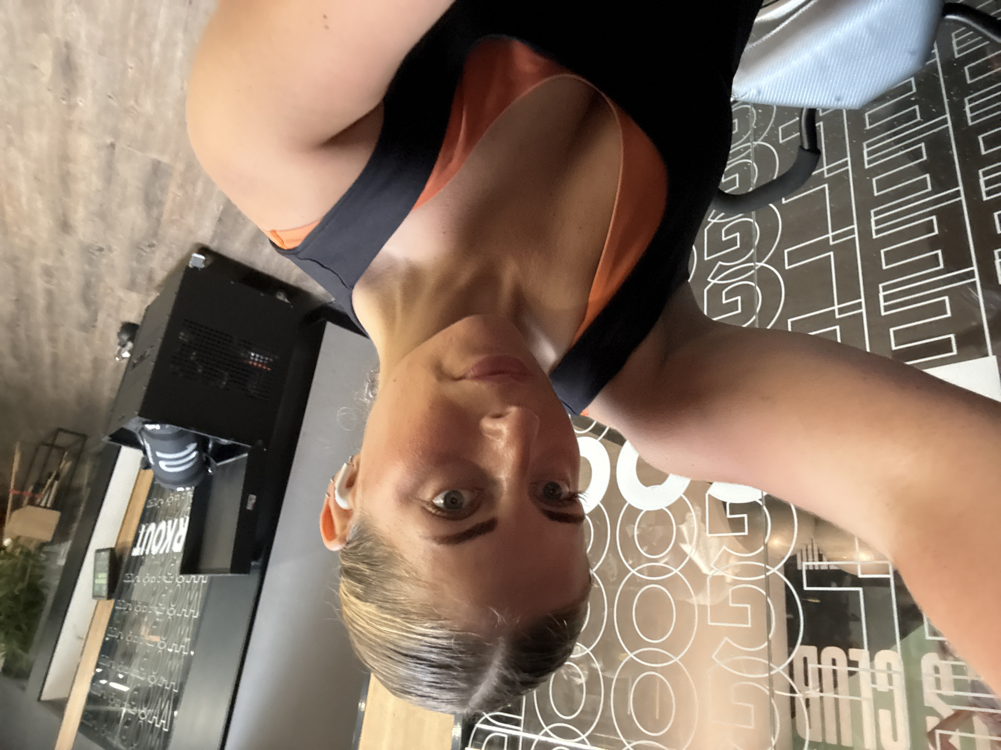 A selfie depicts a blonde woman with an earbud in her left ear, clad in a black and orange sports bra. She is in a gym, as evidenced by the floor's geometric pattern and gym equipment in the background. A machine with the word "RKOUT" visible is in the background. A sign that reads "CLUB 50" can be seen through a glass partition. The location is in Paris, France.