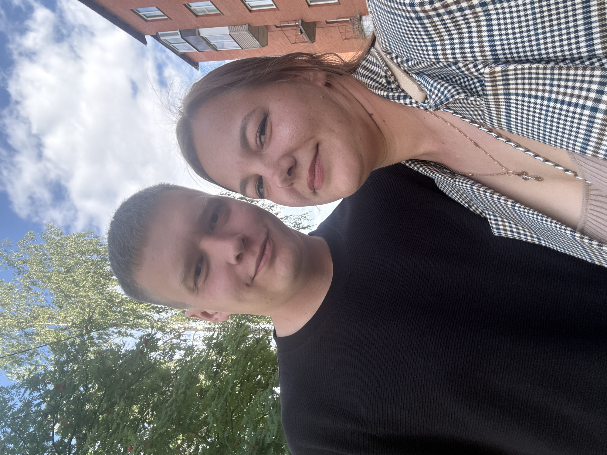 A candid, close-up selfie taken outdoors shows a man and a woman standing close together. The man, on the left, has short brown hair and is wearing a black long-sleeved shirt. He is smiling slightly and looking towards the camera. The woman, on the right, has long brown hair and is wearing a plaid jacket over a light-colored top. She is also smiling and looking at the camera. Behind them, a brick apartment building with balconies is visible, along with trees and a partly cloudy sky. This scene is consistent with a residential area in Dmitrov, Russia.