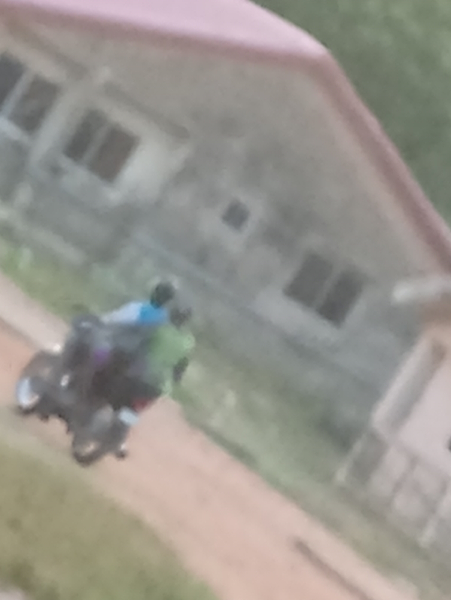 The image depicts two individuals riding a motorcycle on a dirt road. A two-story house stands in the background. The location is Unknown, Unknown.