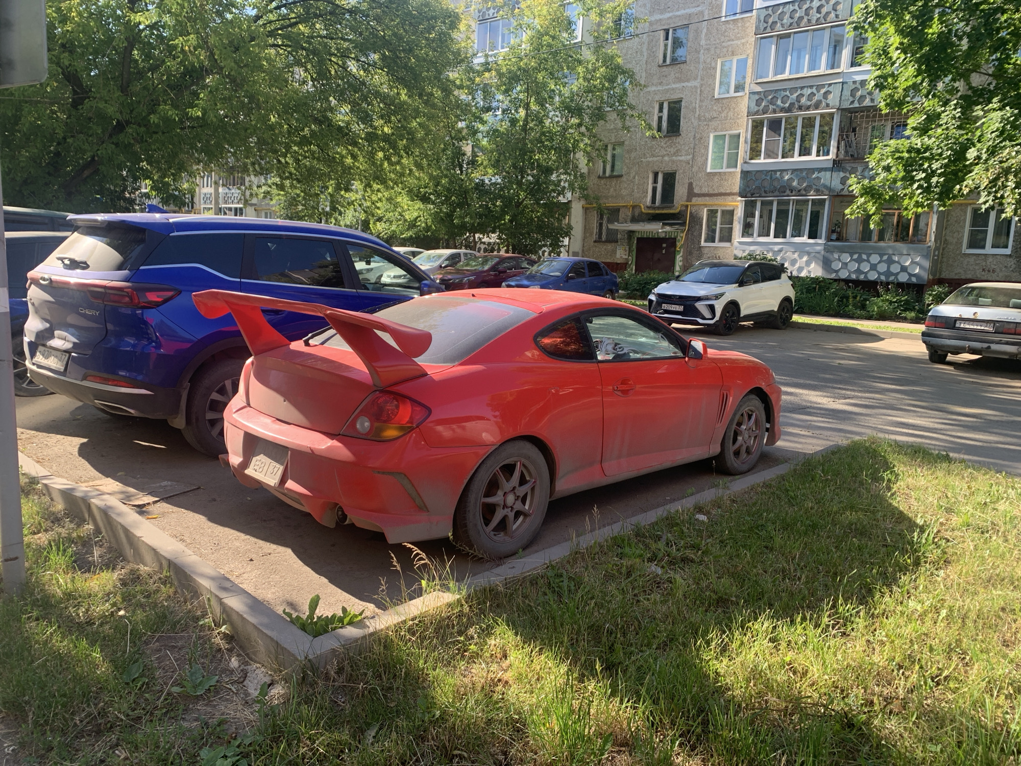 This image captures a sunny daytime scene on a city street in Ivanovo, Russia, likely in a residential area. In the foreground, a bright red sports car with a prominent rear spoiler is parked at an angle. The car appears somewhat dusty. Beside it, a blue SUV is also parked. Further down the street, a white SUV and a light-colored sedan are visible, along with other vehicles partially obscured. The backdrop features a multi-story apartment building with balconies and trees lining the street, suggesting a typical urban environment. The ground is paved with asphalt and there is a strip of grass and a concrete curb separating the parking area from the sidewalk. The overall atmosphere is calm and static, with no people or discernible activity.
