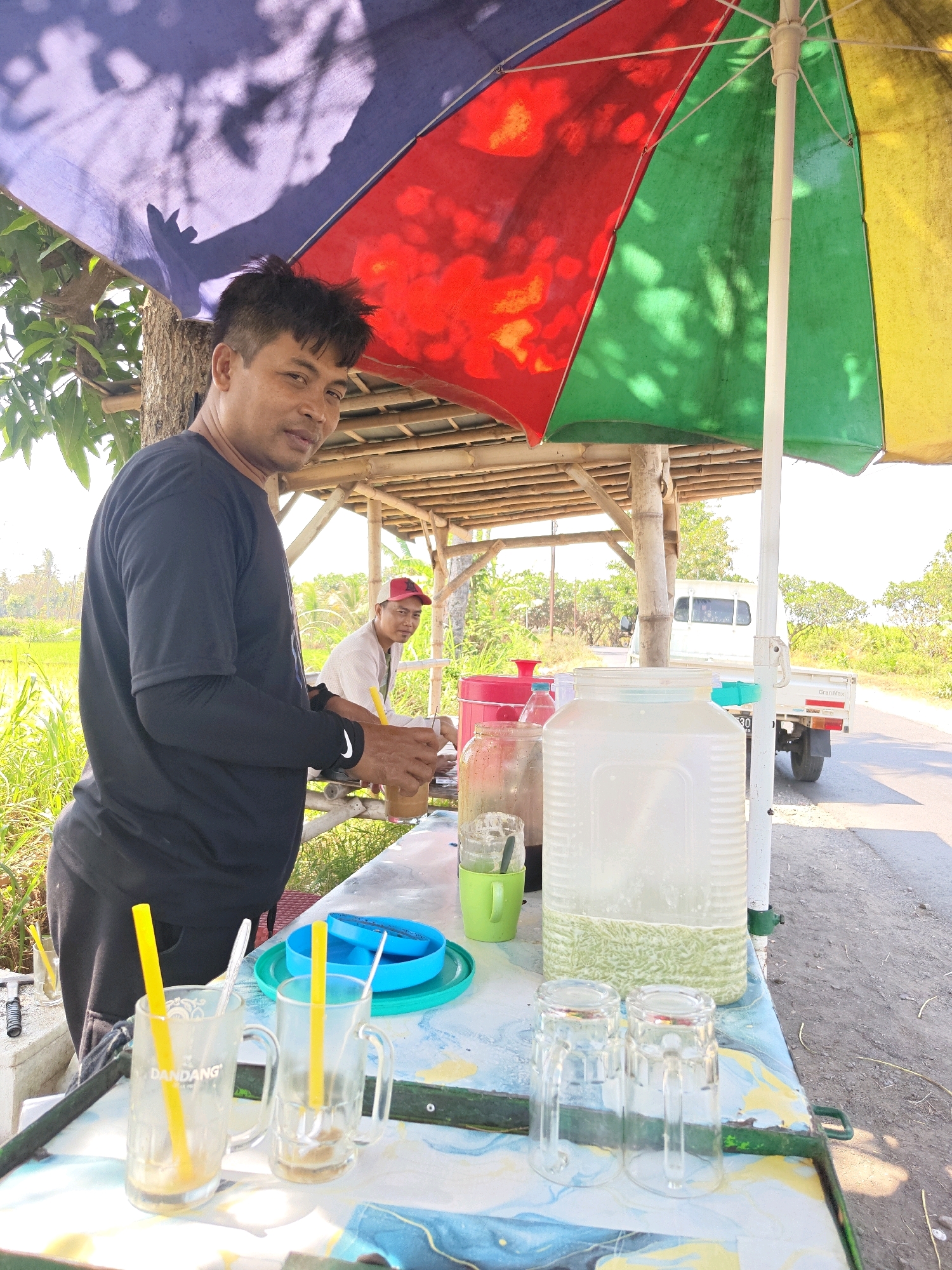 In Klampokan, Indonesia, a man stands behind a table laden with drinks. He is wearing a black t-shirt and dark pants. In front of him, on the table, are several empty glasses, a large plastic jug filled with a light green liquid, and a blue plate. A colorful umbrella, with panels of purple, red, green, and yellow, is open above the scene, casting shadows. In the background, another man in a white shirt and red cap is visible, sitting at a bamboo structure. A white truck is parked on a road next to a field of green grass. The environment appears to be an outdoor roadside stall.