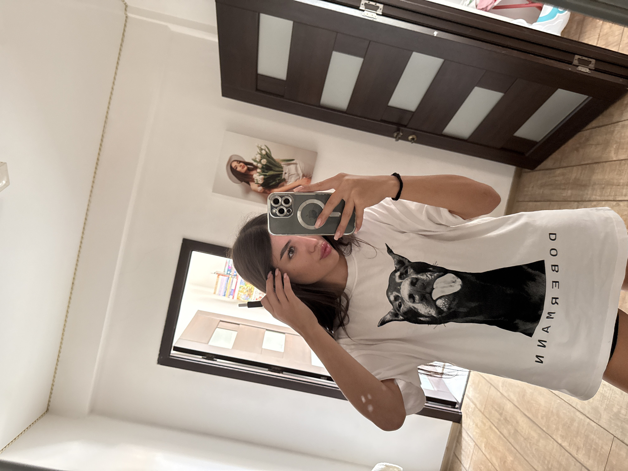 A young woman with long, dark hair is taking a mirror selfie indoors. She is wearing a white t-shirt with a large graphic of a Doberman's head with its tongue out, and the word "DOBERMANN" printed below it in black letters. She is holding a smartphone in her right hand, which is angled to capture the reflection. Her left hand is near her hair, touching it. The background shows a hallway with a light-colored wall, a wooden door on the left, and a dark wooden door with glass panels on the right. There is a framed picture of a woman holding flowers on the wall. The floor is tiled in a wood-grain pattern. The image appears to have been taken in an apartment or house.