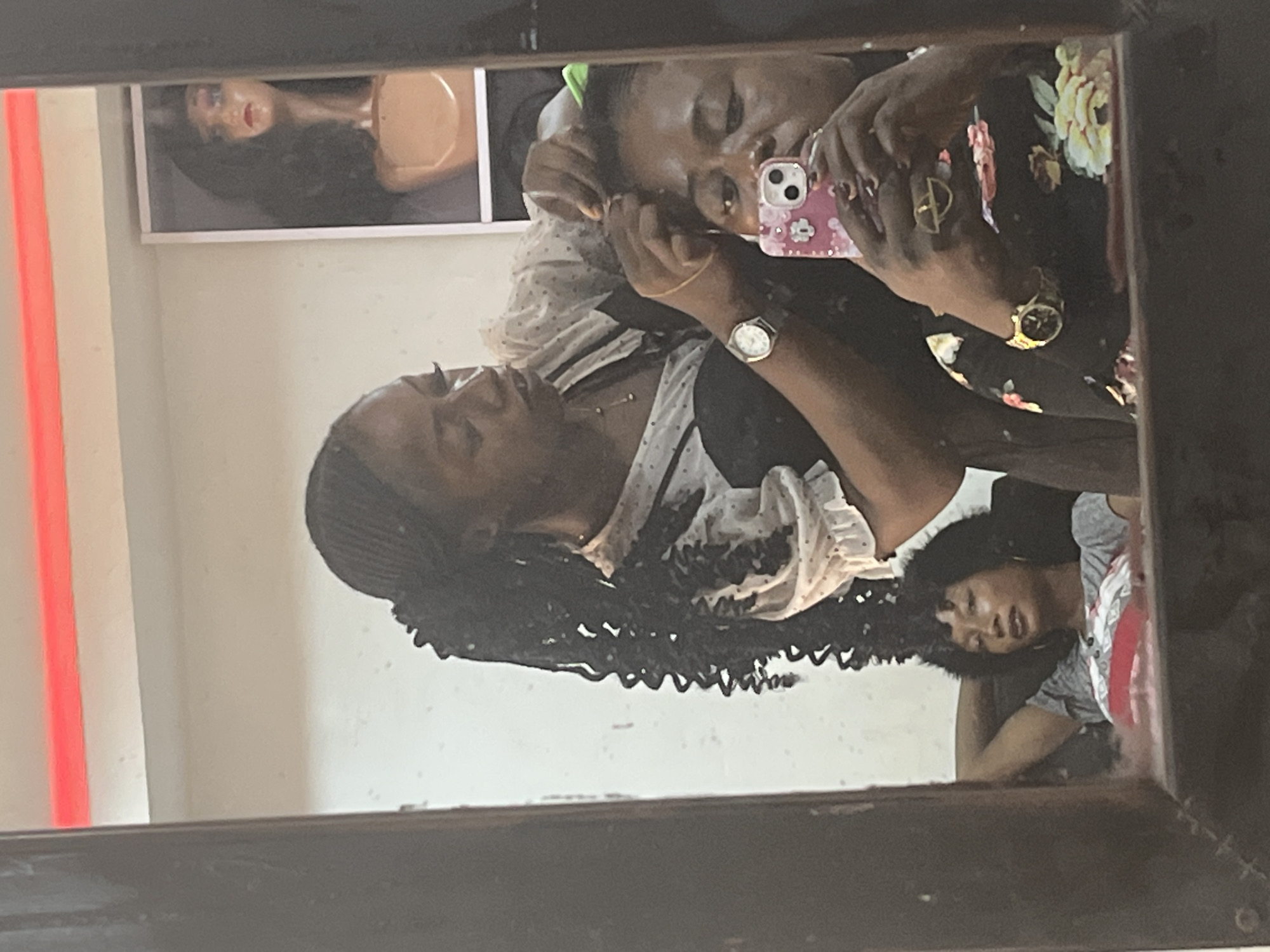 The image captures a reflection within a dark-framed mirror, presenting a scene from a hair salon in Abuja, Nigeria. In the foreground, an adult female client is taking a mirror selfie, holding a pink smartphone with a floral-patterned case in her right hand. She wears a gold wrist watch on her left wrist and a gold ring on her right index finger. Her hair, which is dark, appears to be in the process of being styled, with a green hair tie visible on the crown of her head.
Behind and slightly to the left of the client, an adult female hairdresser is actively working on the client's hair, manipulating strands with both hands. The hairdresser has dark skin, long black braided hair with curly extensions, and is wearing a white, semi-sheer top with polka dots and ruffled short sleeves, layered over a dark, possibly black, garment. She also wears a silver wrist watch on her left wrist and a delicate gold necklace.
In the mid-ground, to the left, a third adult female is observed reclining in a chair, resting her head against her left hand. She has dark skin and short, dark, curly hair, and is wearing a grey t-shirt with a design on the chest and large hoop earrings. Her gaze is directed towards the right side of the frame.
The background includes a light-colored wall with faint speckles. In the upper right quadrant, a mannequin head adorned with a long dark wig is displayed within a white-framed rectangular cabinet. A horizontal red strip, potentially a neon light or decorative trim, spans the upper section of the background wall. The mirror's frame is dark brown or black.