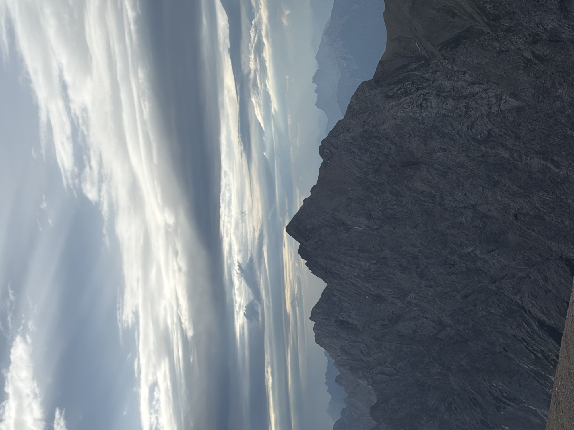 The image shows a majestic mountain range under a dramatic sky. The foreground is dominated by a large, dark, craggy mountain, its surface textured with numerous lines and crevices, suggesting a rocky, rugged terrain. The peak of this mountain is sharp and prominent. Behind it, several layers of smaller, more distant mountains recede into a hazy blue distance. The sky is filled with a complex pattern of clouds. Some are thin and wispy, allowing streaks of sunlight to break through, creating shafts of light that illuminate parts of the sky and possibly the upper reaches of the mountains. Other clouds are thicker and darker, casting shadows and adding depth to the atmospheric scene. The overall impression is one of grandeur and the raw beauty of nature. This scene is likely a view of the Bavarian Alps, possibly from a high vantage point near Garmisch-Partenkirchen, Germany, known for its dramatic mountain landscapes. There are no people or man-made objects visible in the image.