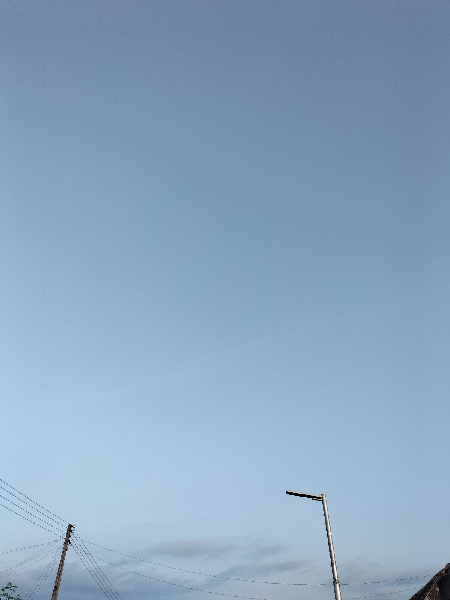 A low-angle, vertical shot captures a clear, pale blue sky dominating the frame. In the lower left quadrant, a wooden utility pole with multiple wires extends upwards, some of which sag downwards. To the right of the utility pole, a modern street lamp with a metallic finish stands tall, its light fixture angled horizontally. In the far lower right corner, a portion of a roofline, possibly from a building, is visible. A thin strip of clouds is present just above the horizon. There are no people or animals visible in the image, and no discernable actions or interactions are taking place. The scene appears to be an outdoor urban or semi-urban environment in Jalingo, Nigeria, characterized by infrastructure elements against a vast sky.