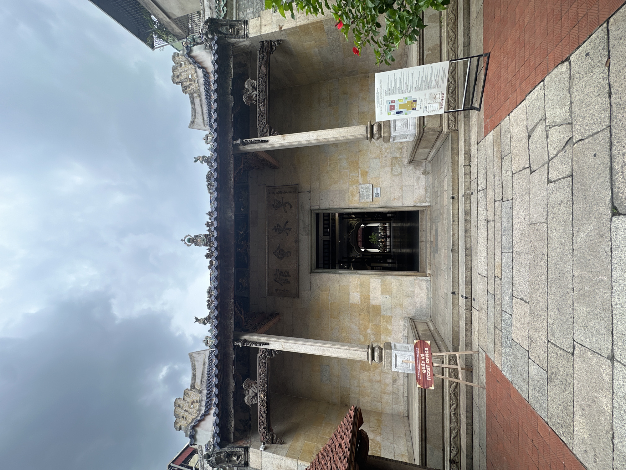 The image shows the entrance to a historical building with traditional Asian architecture in Hanoi, Vietnam. The building's facade is made of light-colored stone blocks, and it features a dark, tiled roof with intricate decorative elements. Above the entrance, a wooden plaque with Chinese characters is mounted. The entrance itself is an open doorway, leading into a dimly lit interior.
In front of the building, a stone pathway is laid out, with a section of red patterned tiles on the left side. Two signs are placed on the pathway. One sign, on a wooden easel, reads "QUẦY VÉ TICKET OFFICE" in red lettering. Another larger sign on a black stand displays a map and information in both Vietnamese and English.
The surrounding environment appears to be overcast, with a cloudy sky visible at the top of the image. There is some greenery, including a bush with red flowers, on the right side of the frame. The overall scene depicts a cultural or historical site, likely a museum or temple, with clear signage for visitors.