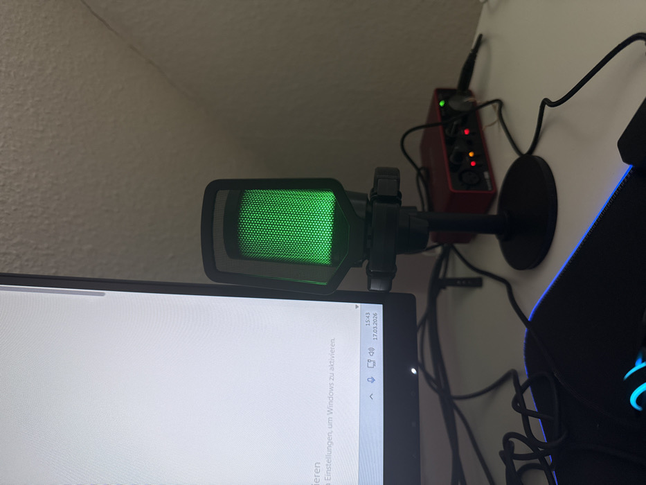 This image depicts a segment of a computer workstation located in Vellmar, Germany. The foreground features a white desk surface. On the left, a vertical computer monitor displays a white screen with German text at the bottom, partially visible as "Einstellungen, um Windows zu aktivieren." The Windows taskbar is visible, showing the current time as 15:43 and the date as 17.03.2026.
To the right of the monitor, a black cylindrical USB microphone with a prominent green illuminated mesh grill stands on a desk mount. Behind and to the right of the microphone, a red rectangular Focusrite Scarlett 2i2 audio interface is visible, powered on with red and orange/yellow indicator lights active. Various black cables are scattered across the desk surface, connecting the devices. A section of a black mousepad with blue LED edge lighting is visible in the lower-left corner. The background consists of a light-colored, textured wall.