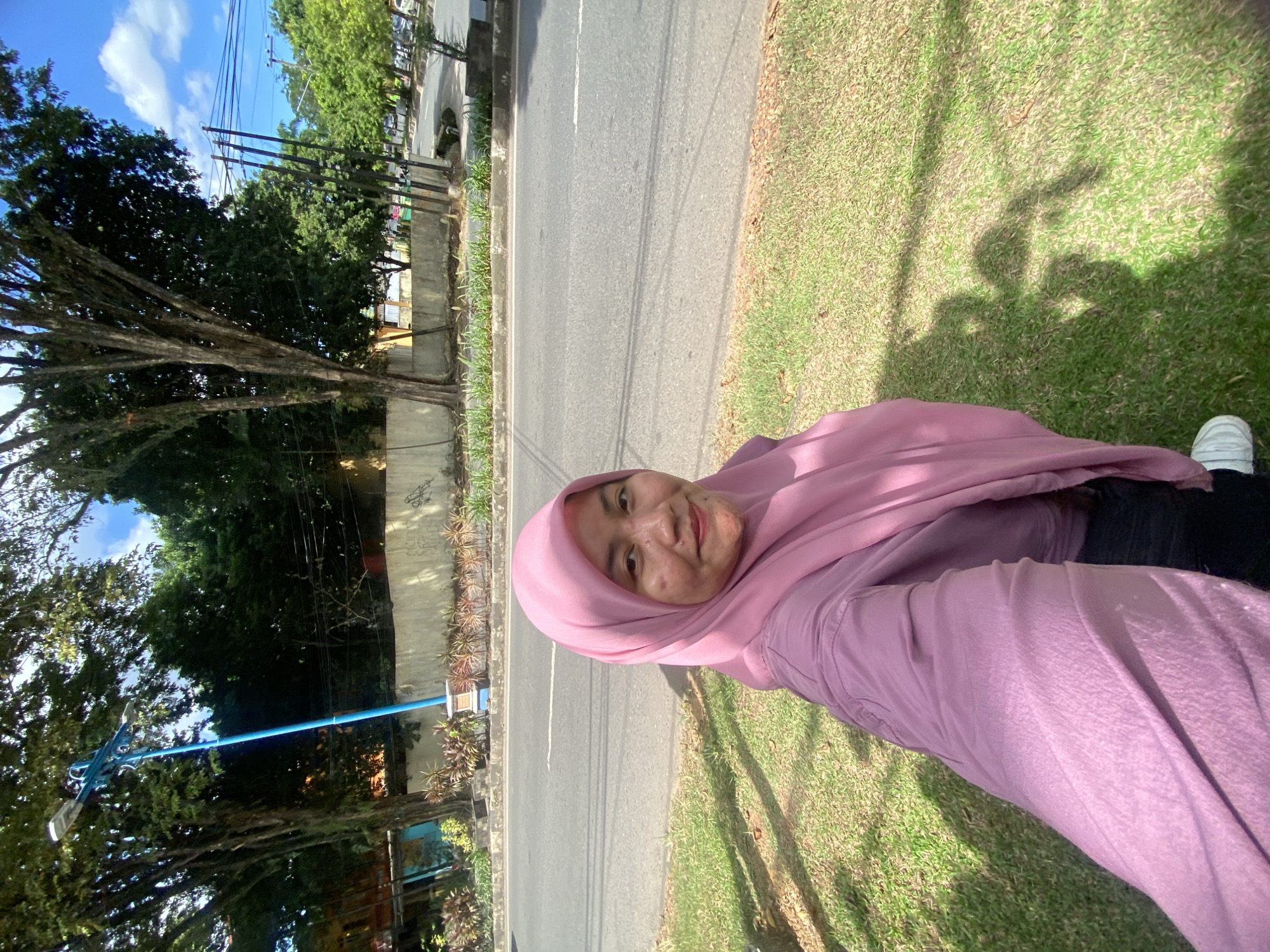 A woman wearing a pink hijab and a pink shirt is taking a selfie in a grassy area next to a road in Banjarbaru, Indonesia. The road has multiple lanes and is bordered by a concrete wall with some plants. In the background, there are trees, utility poles, and buildings. The sky is blue with some white clouds. The woman's shadow is cast on the grass.