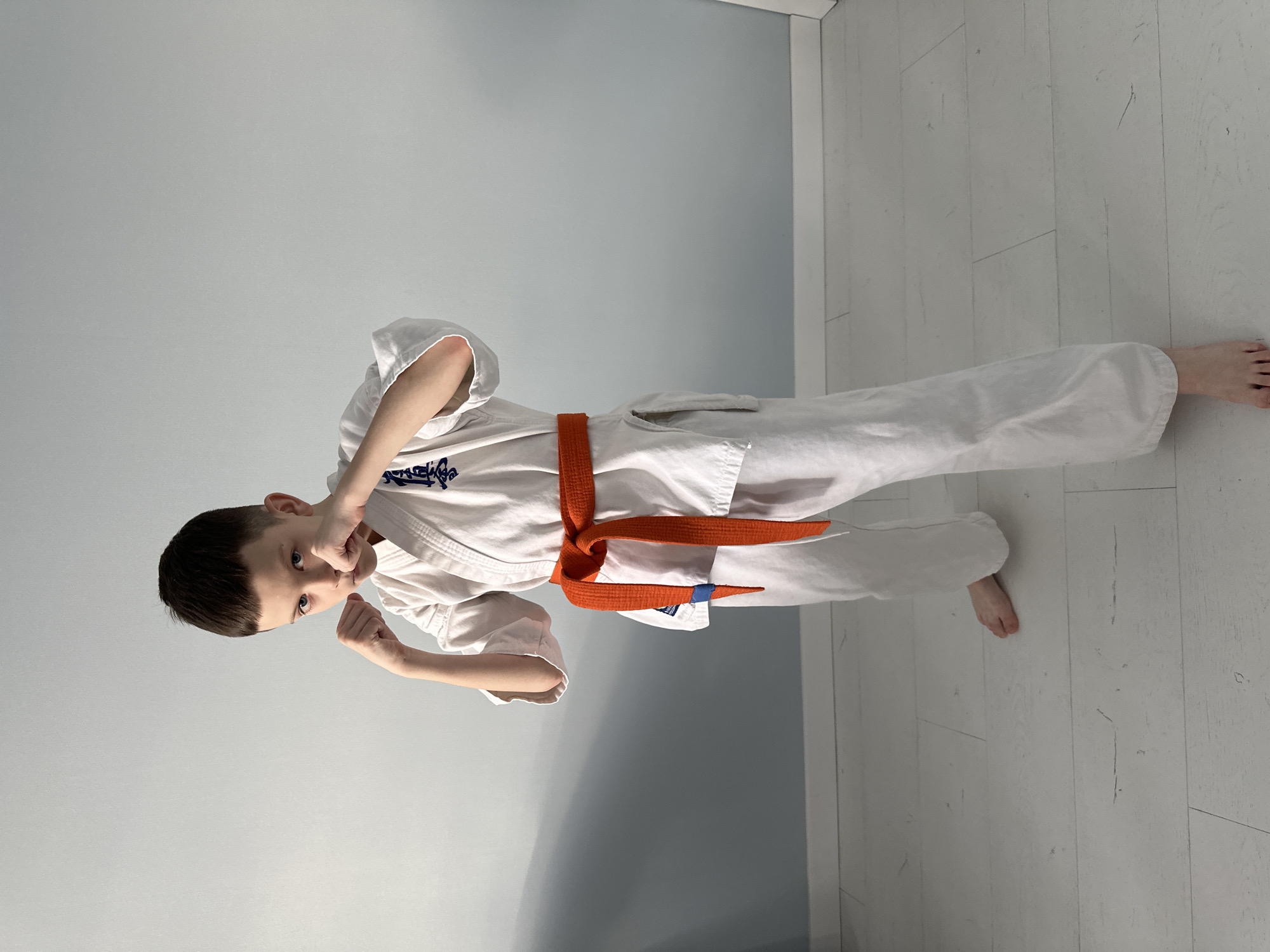 A young boy, appearing to be of primary school age, is captured standing in a dynamic martial arts pose in an indoor setting in Прикамский, Russia. He is dressed in a crisp white karate gi (uniform) cinched with a bright orange belt. His fists are clenched and raised in front of him, suggesting a fighting or defensive stance. He looks directly at the camera with an intense yet seemingly playful expression, eyes wide and focused. His dark hair is neatly cut short.
The background features a plain, light blue-gray wall that dominates the upper and middle portions of the image. The lower part of the frame reveals a floor made of light-colored wooden planks. The lighting is soft and even, indicating daytime conditions. The boy is barefoot. On the left chest area of his gi, there is a dark blue emblem or text, likely Japanese kanji, which is partially visible but not clearly legible. The scene conveys a sense of youthful energy and martial arts discipline.