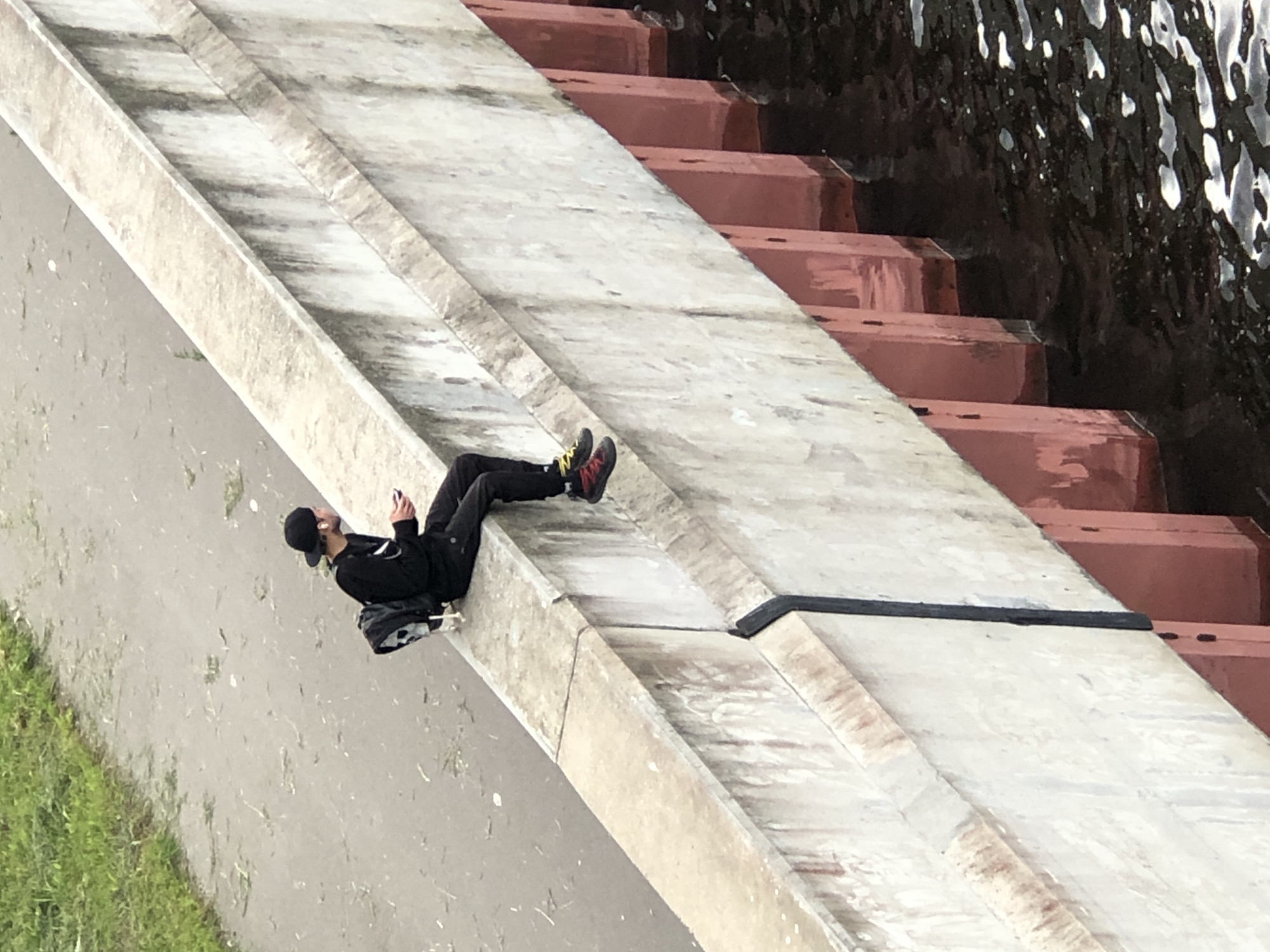 A young person wearing a black baseball cap and black clothing, including a backpack, sits on the edge of a concrete structure. The person is holding a mobile device and appears to be listening to something through earbuds. The structure overlooks dark, rippling water with several vertical red-colored supports visible beneath the concrete. The ground beside the concrete structure is a mix of grey pavement and green grass.