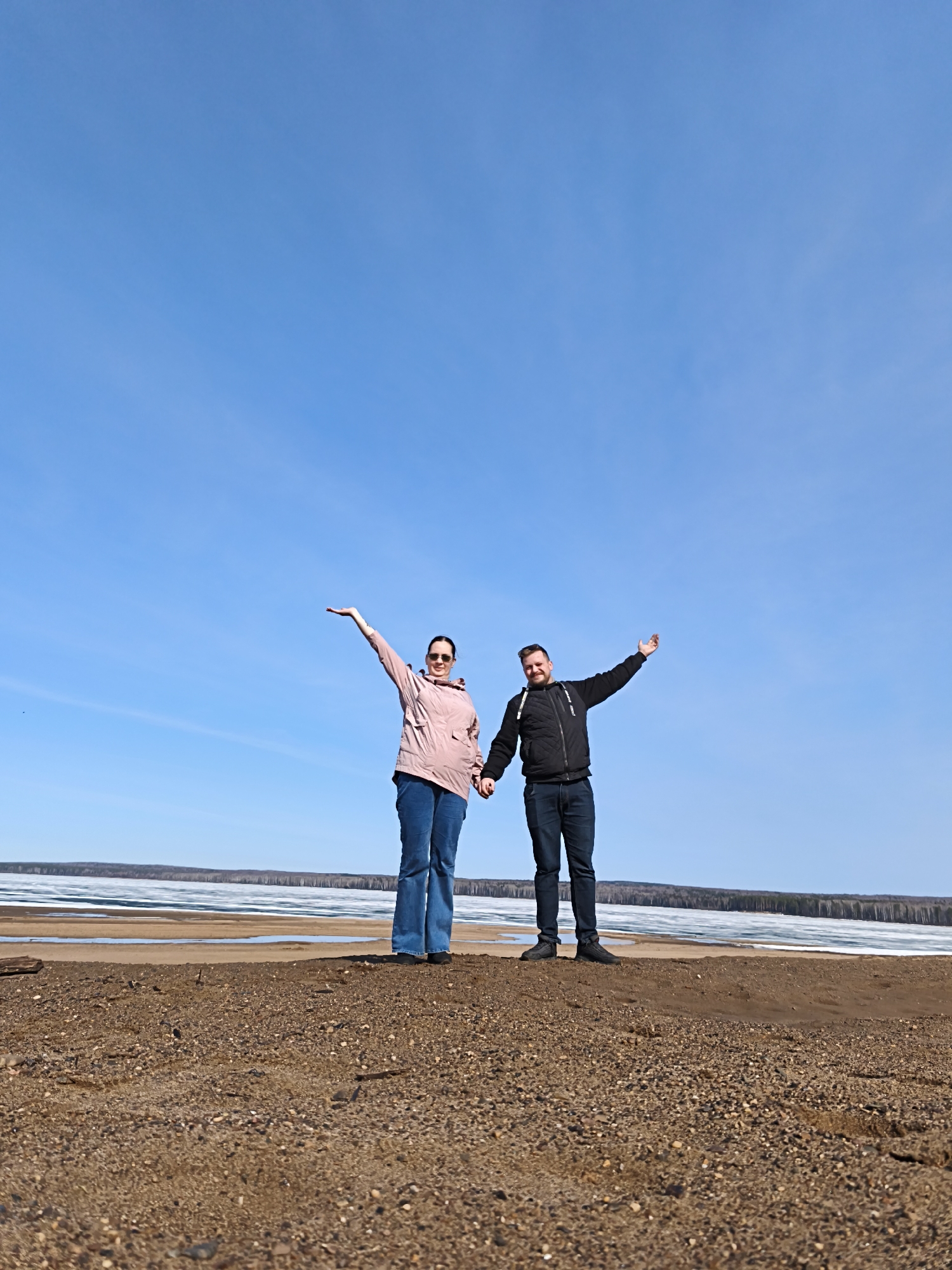 A full shot depicts a man and a woman standing hand-in-hand on a sandy terrain under a clear blue sky. The woman, positioned on the left, wears a light pink jacket, blue jeans, and sunglasses, and has her left arm raised. The man, standing on the right, is dressed in a black jacket and dark jeans, with his right arm also raised. Behind them, a body of water with patches of ice is visible, bordered by a line of trees in the distance. The foreground consists of a sandy surface with small rocks.