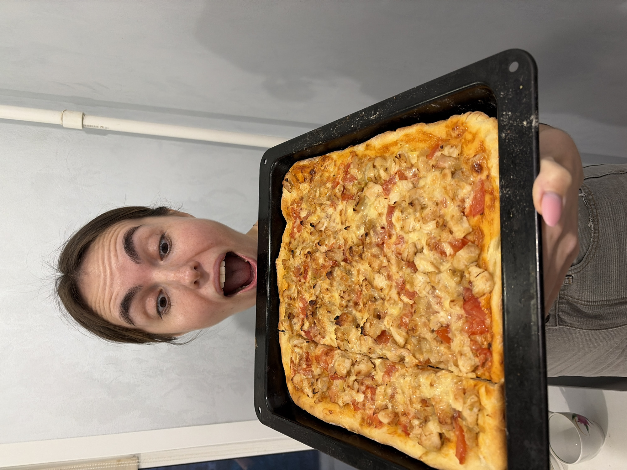 A person is holding a rectangular pizza on a black baking sheet at an angle. The person has brown hair and is wearing a grey shirt. The pizza appears to be cut into two pieces and has a topping of cheese, tomatoes, and chicken. The background consists of a white wall with a white pipe running across it. A mug can be seen on a white surface in the lower right corner of the image. It is not possible to determine the location is Sarapul, Russia from the image.