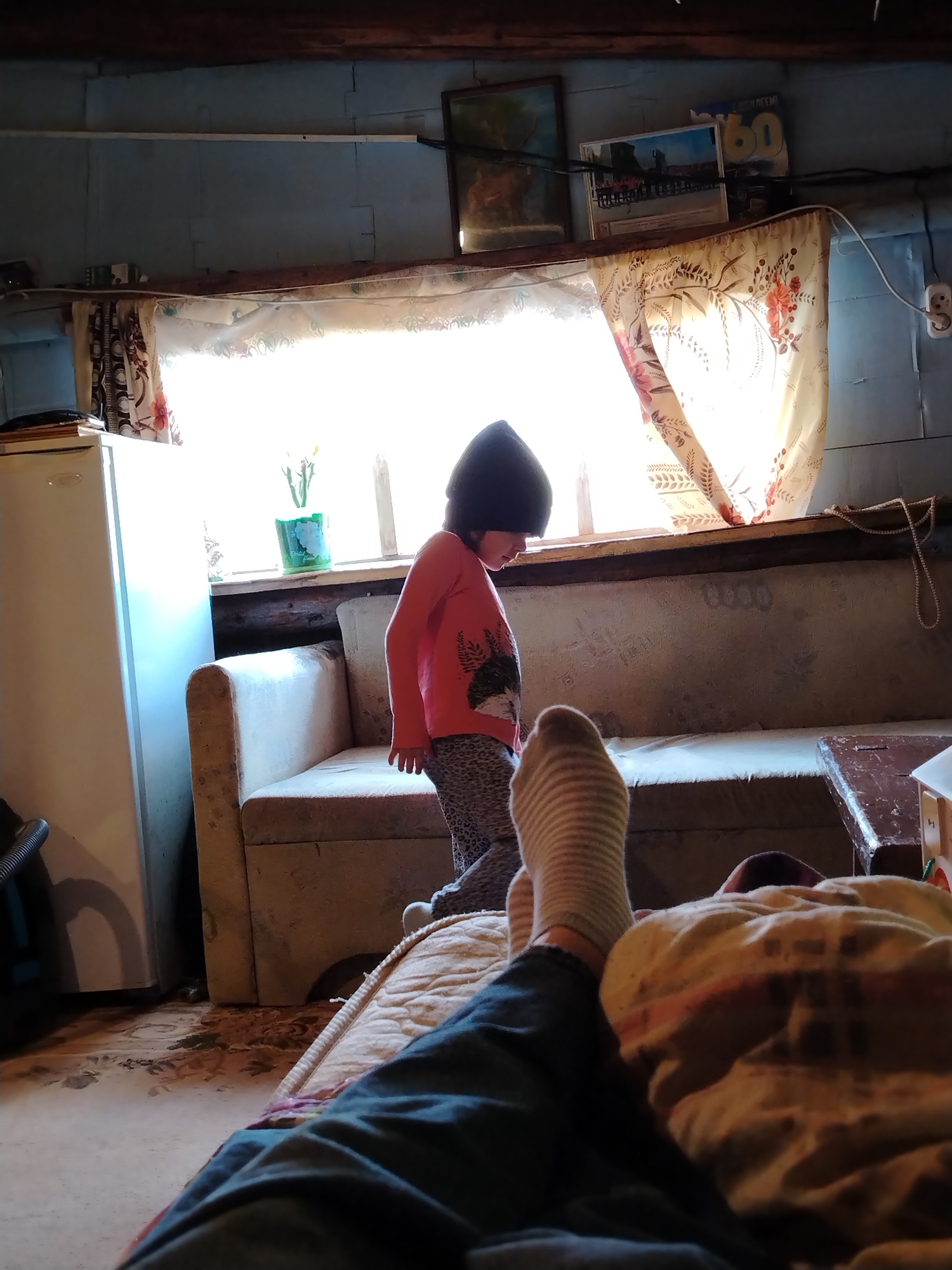 A photograph taken in what appears to be a residential interior, possibly in the city of Гожан, Russia, shows a room with light blue planked walls and a dark wood beam ceiling. A single window is draped with patterned curtains. A young child, wearing a dark beanie, a red shirt, and patterned leggings, stands near the window. A sofa is positioned below the window. A white refrigerator is to the left of the sofa. A person's legs and feet, covered with socks and denim, are extended into the foreground from a horizontal position, as if reclining on a bed or another seating arrangement. The foreground also includes a patterned blanket. Above the window, several framed pictures and a calendar are placed on a shelf. The lighting in the room is somewhat dim, and the overall impression is one of a rustic or older home.