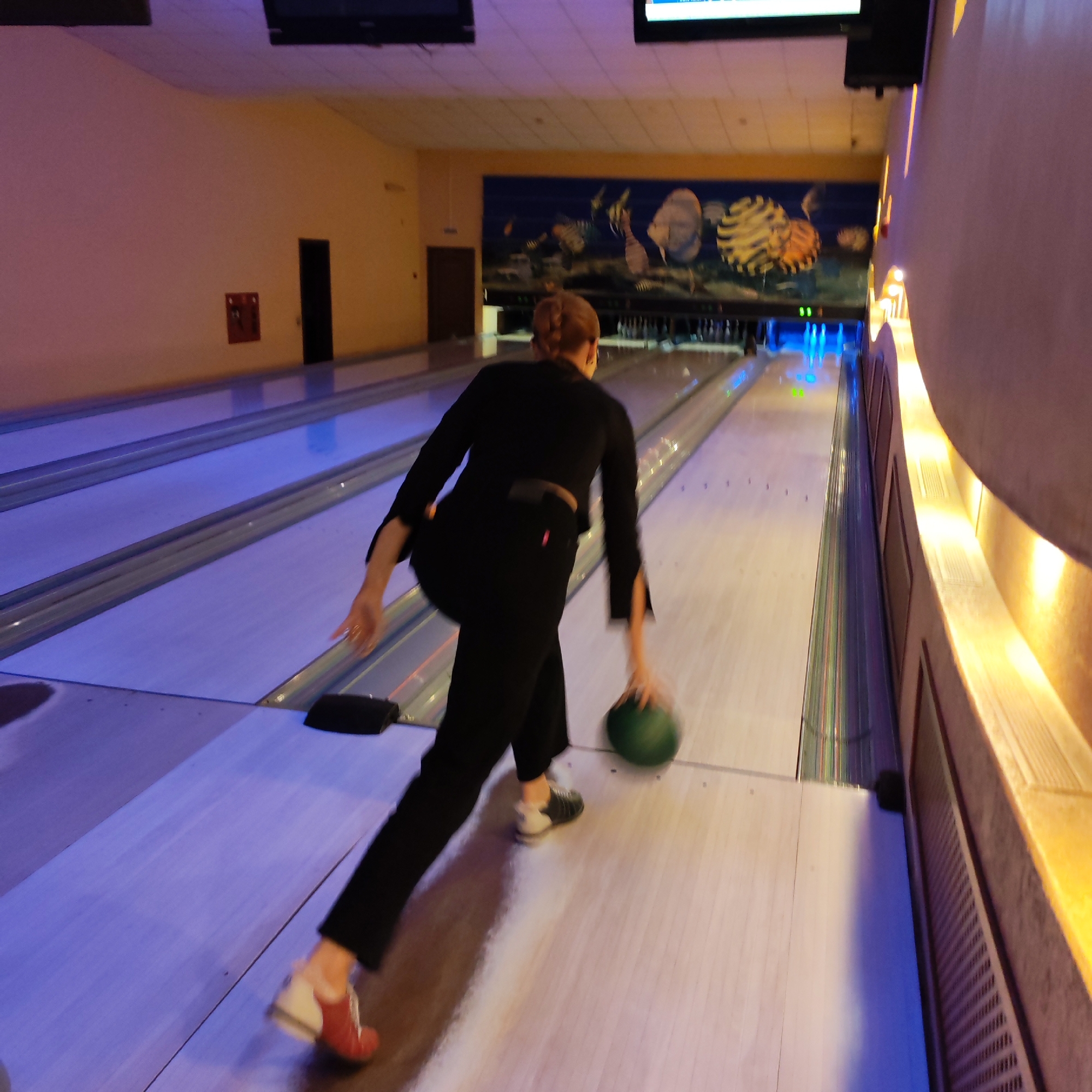 An overhead-angled photograph depicts a bowling alley in Chaikovsky, Russia, from behind a person in the act of bowling. A person with auburn hair, styled in a bun, is dressed in a black long-sleeved top and black trousers. They are captured in motion, leaning forward and extending their right arm to release a green bowling ball onto a lane. The person wears two distinct bowling shoes: a white and black shoe on their left foot and a red and white shoe on their right foot.
Two bowling lanes are visible, illuminated by blue and purple neon lighting along their gutters and approach areas. The lanes themselves are light-colored, resembling polished wood. At the far end of the right lane, white bowling pins are arranged, with some visible in the pinsetter area, which emits blue light. Beyond the pins, a large mural covers the wall, depicting stylized, abstract forms in blue, yellow, and orange hues. Two dark rectangular screens, likely for scoring, are mounted high above the lanes.
To the left of the lanes, a light peach-colored wall features a dark rectangular door and a red wall-mounted fire extinguisher box. To the right, a parallel wall with light-colored panels and horizontal ventilation slats is illuminated by warm, recessed lighting elements, casting an orange glow. The ceiling is light-colored and textured.