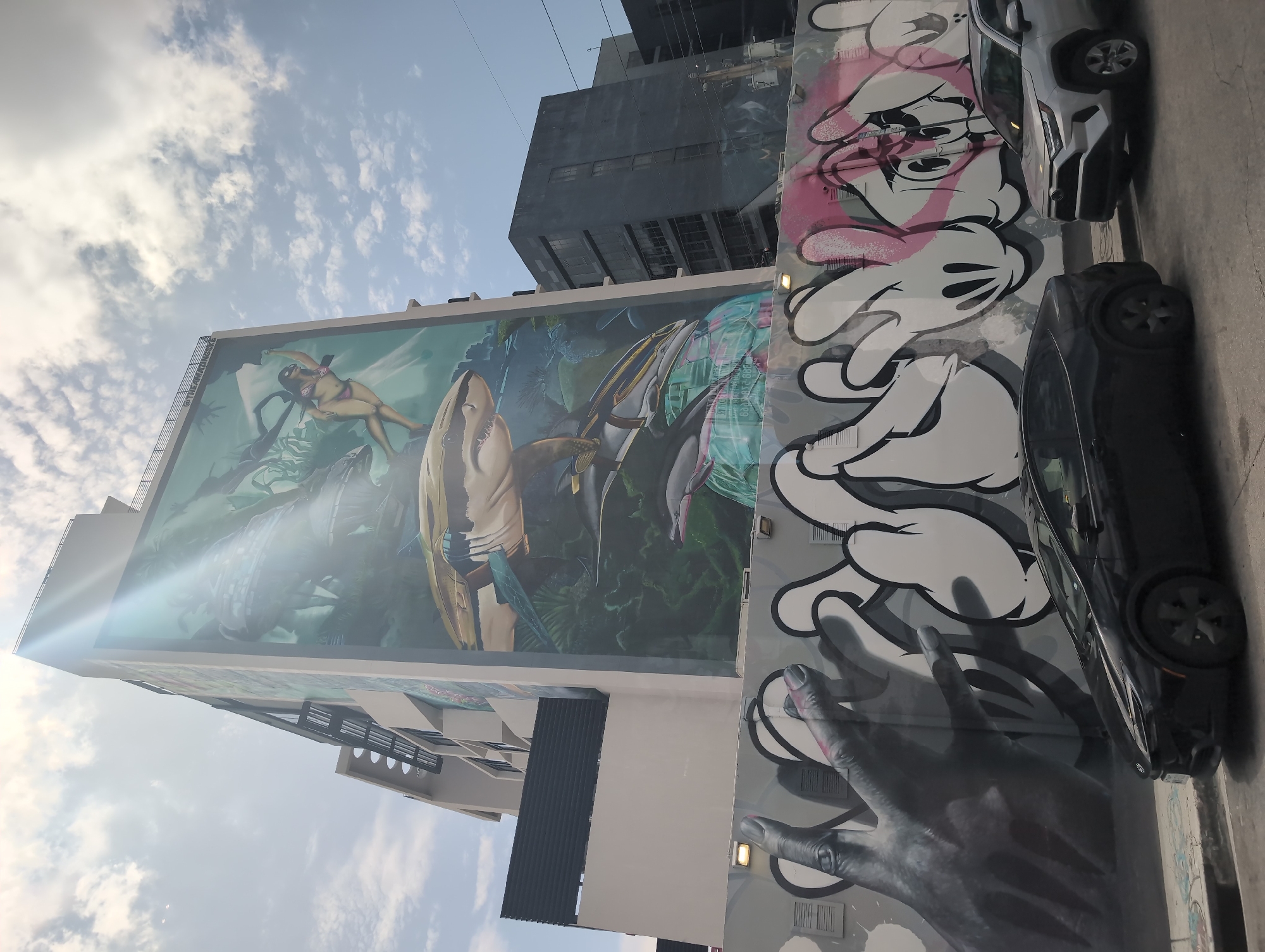 The photograph captures a mural on the side of a multi-story building in Miami, Florida. The mural depicts an underwater scene with a shark, dolphins, and a person in a bikini swimming. The building has a modern architectural style with balconies and windows. In front of the building, a graffiti-covered wall features stylized hands and cartoon characters. Two cars, a black sedan and a silver SUV, are parked on the street in front of the graffiti wall. The sky is partly cloudy, and sunlight is visible, creating a lens flare effect. The overall scene is vibrant and urban, showcasing street art and modern architecture.
