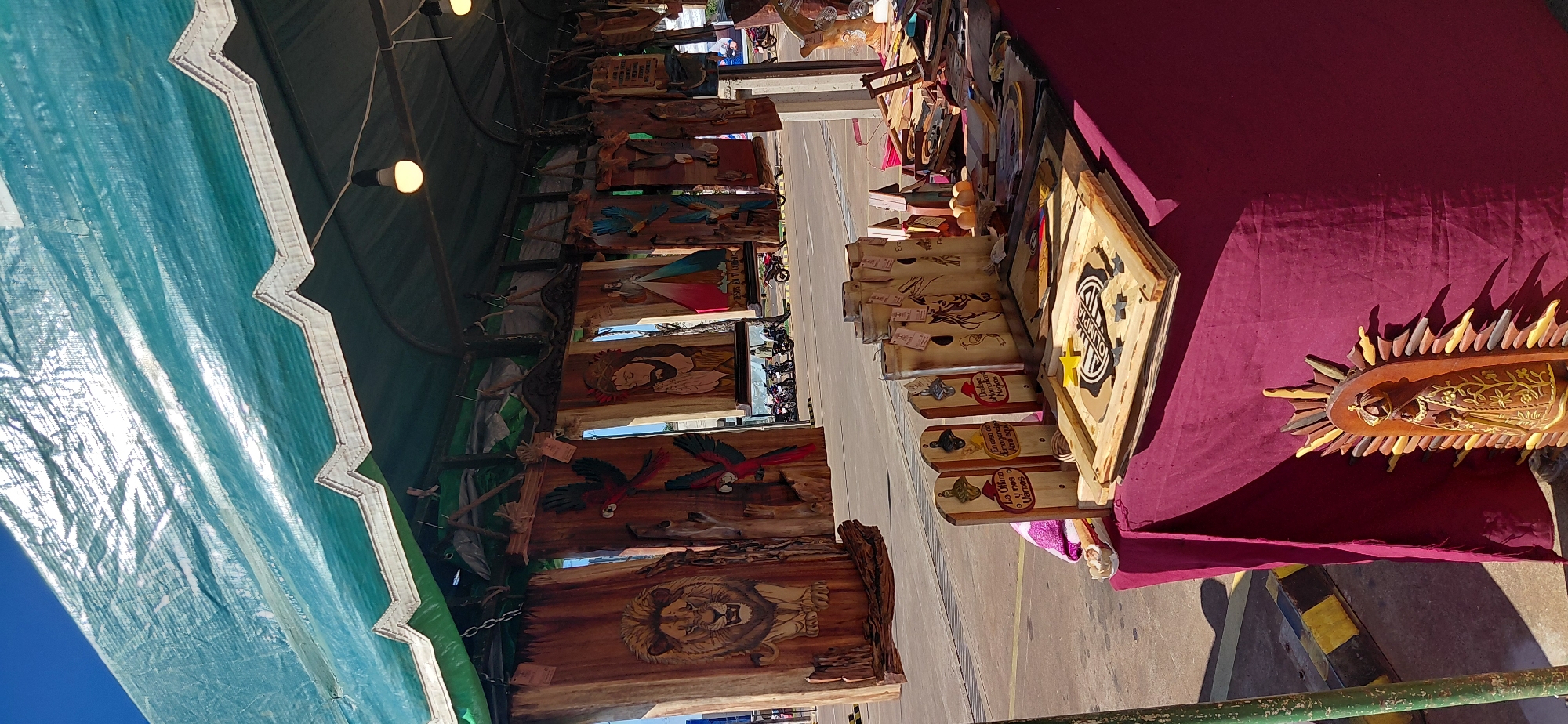 This is a photograph of an outdoor market stall in Encarnación, Paraguay. The stall is covered by a green tarp that is supported by a metal frame. Underneath the tarp, a variety of wooden crafts are displayed. In the foreground, a table covered with a dark red cloth showcases several items, including wooden bottle openers with inscriptions, birdhouses adorned with carved animals, and a framed wooden piece with a logo that reads "OLIMPIA." To the left of the table, a wooden statue of the Virgin Mary is visible.
Behind the table, hanging from the stall's structure, are large wooden panels carved with various designs. These include a lion's head, parrots, and religious imagery, such as depictions of Jesus and other figures. In the background, a street with parked motorcycles and people is visible, suggesting a public area. The overall impression is that of a lively market scene with artisanal products for sale.