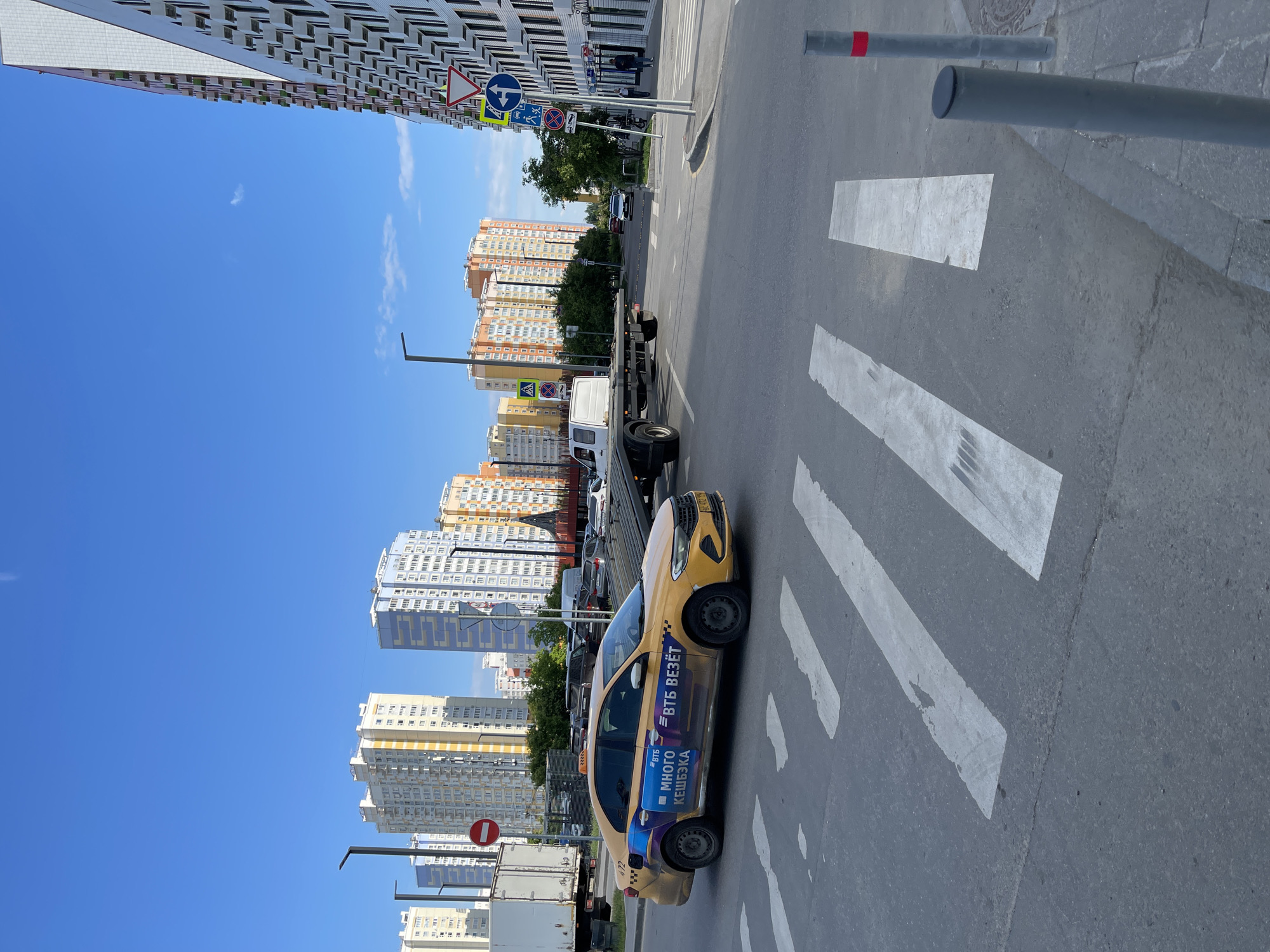The image presents an urban street scene in Kommunarka, Russia, captured on a bright, clear day under a vivid blue sky with minimal cloud cover. The primary subject is a yellow taxi cab, positioned on an asphalt road at a pedestrian crosswalk marked by bold white stripes. The taxi features prominent branding on its side, displaying "ВТБ ВЕЗЁТ" (VTB VEZYOT) and "МНОГО КЕШБЭКА" (MNOGO KESHBEKA), along with the number "472".
In the background, a busy urban environment unfolds with numerous multi-story residential buildings. To the left, a tall, modern building with a repeating geometric pattern on its facade stands out. Across the street and further down, an array of other high-rise apartments in various colors like orange, yellow, and blue define the cityscape, suggesting a densely populated area. Green trees are scattered throughout the scene, adding natural elements to the concrete jungle.
Traffic infrastructure is visible, including a white flatbed truck or tow truck behind the taxi, and other cars in the distance. Several street signs are present, notably a pedestrian crossing sign, a blue circular sign with a white arrow indicating direction, and a red circular "No Entry" sign. Street lamps and grey bollards line the sidewalks, which are made of concrete pavers. The scene depicts a typical, active city street during daylight hours, showcasing a mix of modern architecture and daily urban life.