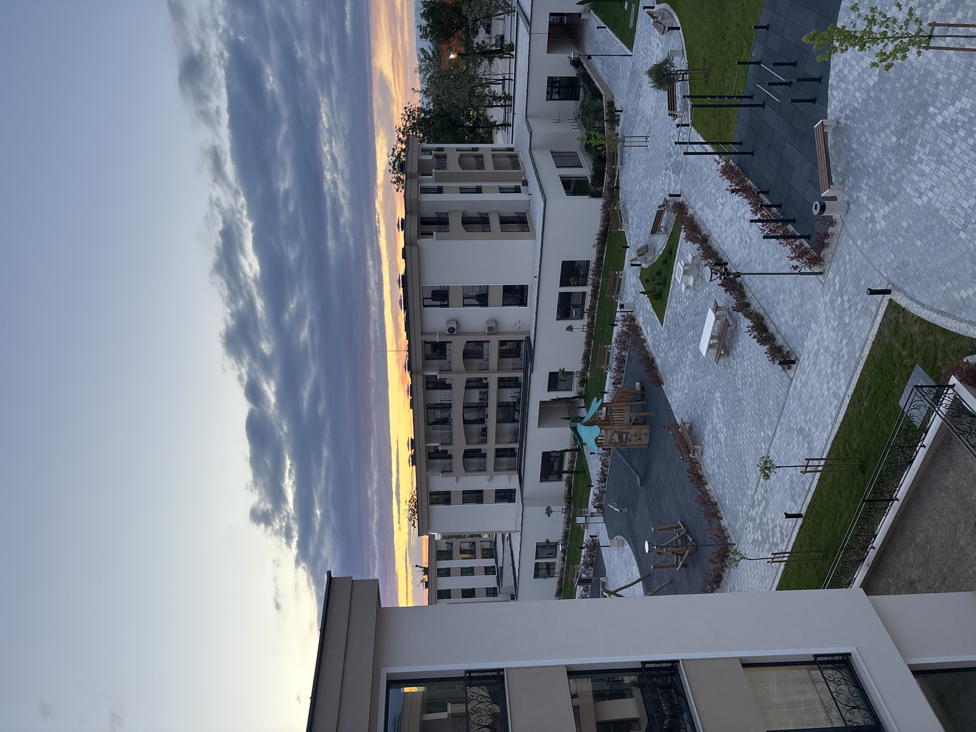 An elevated view in Plovdiv, Bulgaria shows a modern residential complex at sunset. Multiple white apartment buildings with balconies and visible air conditioning units border a paved courtyard. A playground with wooden structures is visible in the courtyard. Exercise equipment, benches, and planted greenery are also present. The sky displays hues of gray, purple, and orange clouds as the sun sets. A foreground building section with windows and decorative ironwork is included in the bottom of the frame.