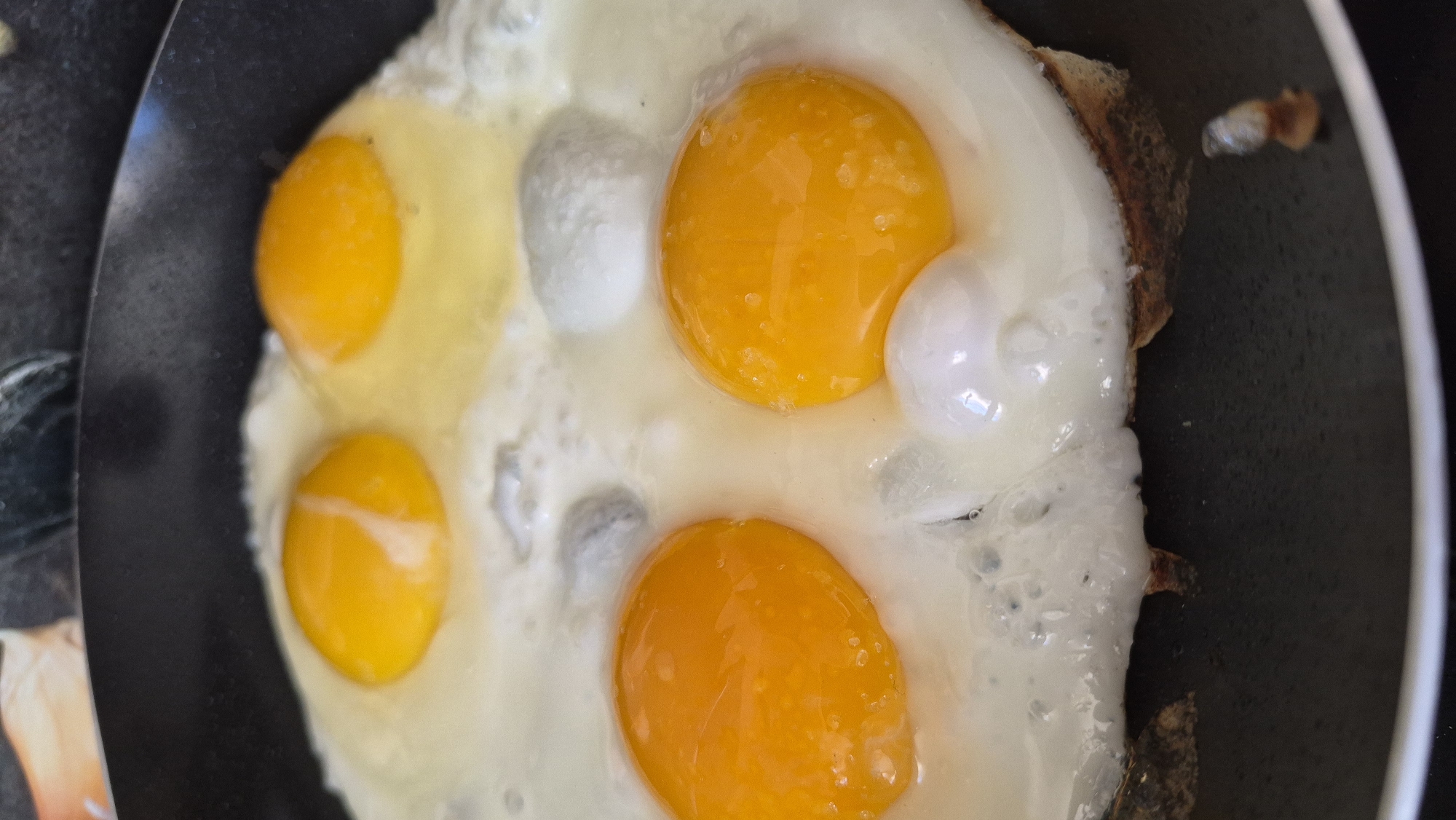 An overhead, close-up shot shows four fried eggs in a black frying pan. The eggs are arranged in a square formation, with two eggs in the front and two in the back. The yolks of the front two eggs are bright orange and appear to be runny. The back two eggs have lighter yellow yolks and are slightly more cooked. The whites of the eggs are opaque and slightly bubbly. The pan itself appears to be well-seasoned, with some browning on the edges. The background is blurred, but it appears to be a kitchen counter with some indistinct objects. There is no indication of Krauja, Latvia in the image.