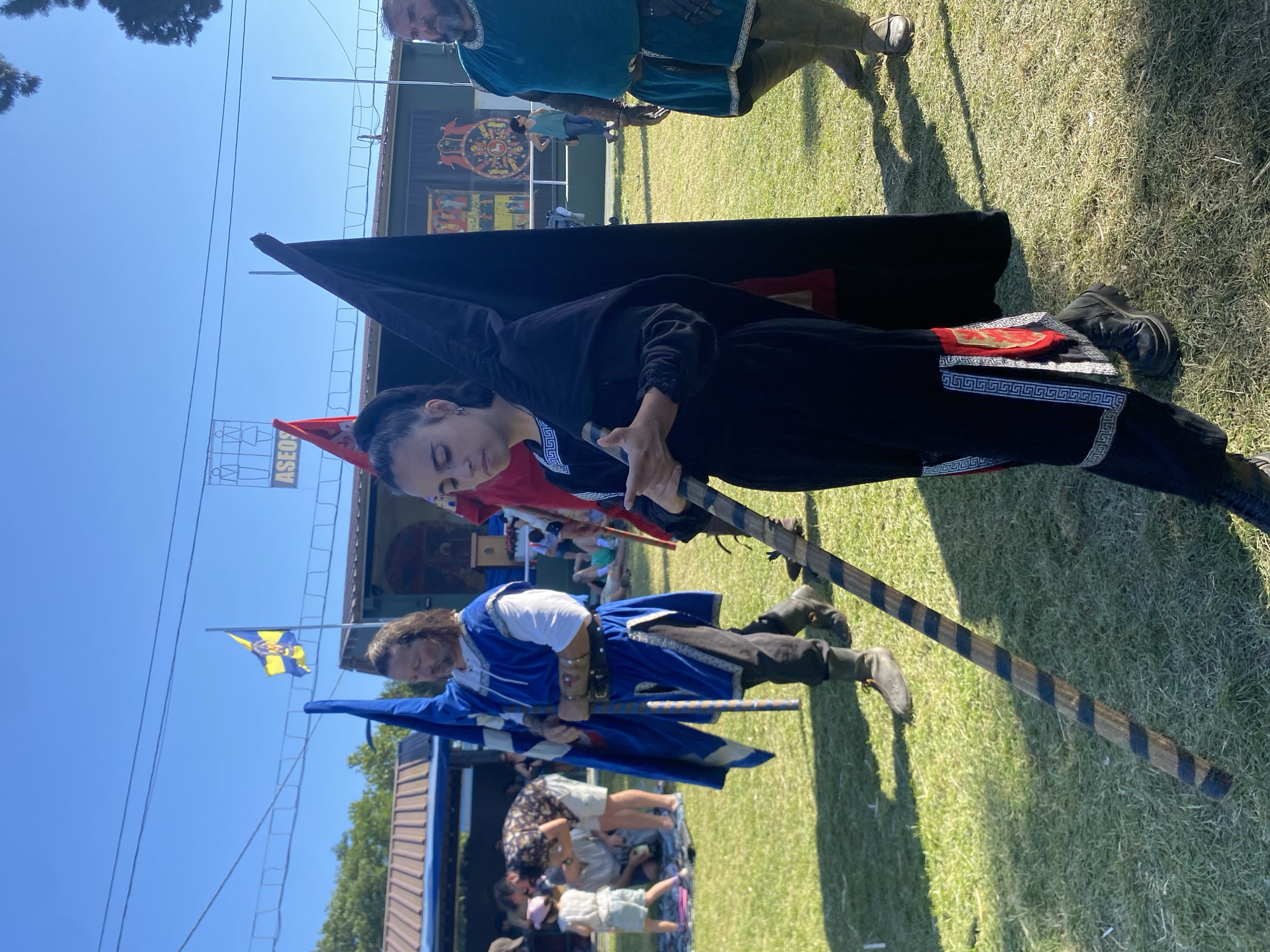 In Llanera, Spain, on a sunny day, a group of people in medieval-style attire are participating in an outdoor event. In the foreground, a young woman in a black hooded cloak with ornate trim is holding a black flag and a wooden staff. To her left, a man with long brown hair, dressed in a blue tunic and white shirt, carries a blue and yellow flag. Behind them, a man in a teal tunic is visible. The background shows a grassy field, tents, and spectators. The sky is clear and blue.
