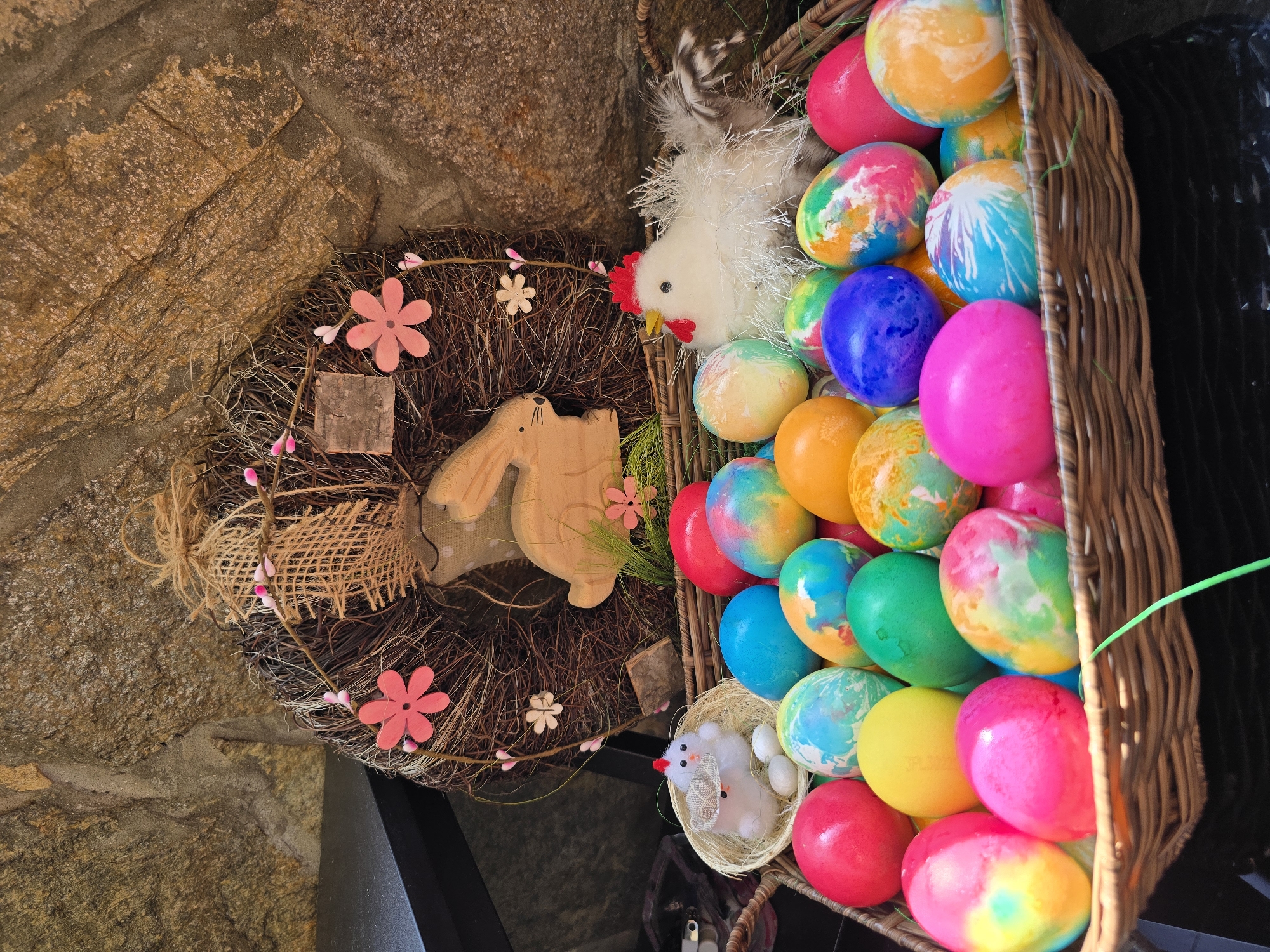 The image depicts an Easter-themed scene, likely in a retail or public space. A wicker basket overflowing with brightly colored Easter eggs in various shades of pink, red, orange, blue, and green dominates the right side of the frame. Nestled among the eggs are two small, white, plush animal figures: a chicken and a mouse. To the left of the basket, a decorative wreath made of twigs, burlap, and wooden adornments, including pink flowers and a rabbit-shaped cutout, rests against a textured stone wall. The stone wall suggests an exterior setting or a rustic interior. The image hints at the festive Easter season and the decorative displays associated with it. It is impossible to confirm the image location is Burgas, Bulgaria with only the visual information provided.