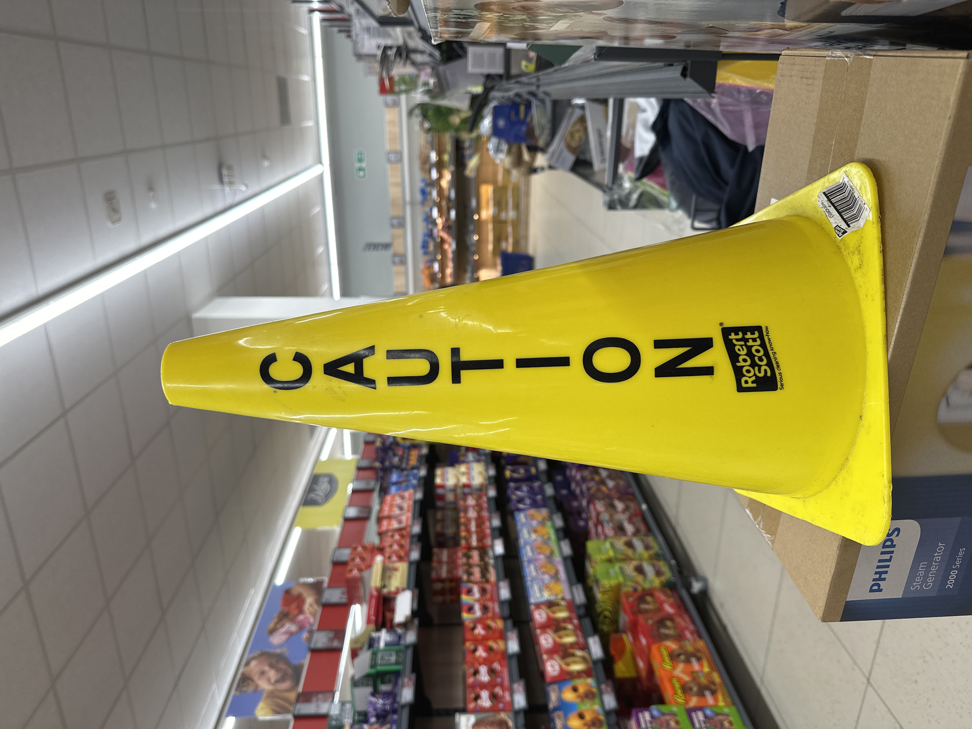 A bright yellow "CAUTION" cone, displaying the vertically printed word "CAUTION" in black capital letters and a "Robert Scott Serious cleaning know-how" logo, is positioned centrally in the foreground. A barcode sticker is present on its base. The cone rests on a brown cardboard box, partially revealing "PHILIPS Steam Generator 2000 Series" text. The scene is an interior view of a supermarket aisle in London, United Kingdom. To the left, multiple shelves are fully stocked with various packaged confectionery and snack items, with promotional banners including one featuring a smiling individual and another with a "Deluxe" logo suspended above. The background shows further supermarket aisles with indistinct products, a possible bakery section, and a blue shopping basket on the floor. The ceiling is a white tiled grid with numerous linear fluorescent lights. An emergency exit sign is visible high on a distant wall. The flooring is a light-colored tiled surface. No individuals are present within the frame. The "Deluxe" branding suggests an Aldi supermarket.
