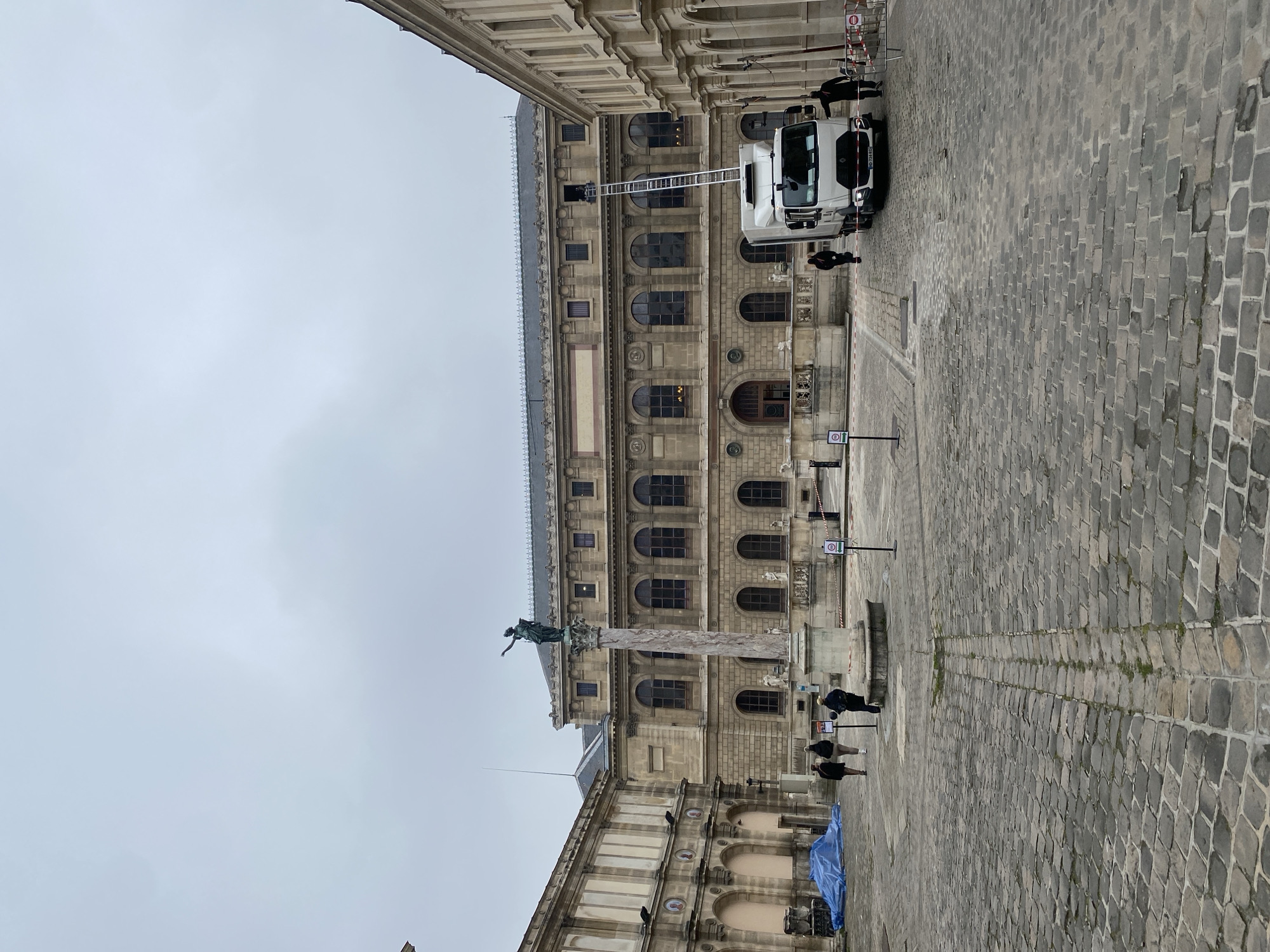 The image shows a scene in Paris, France, featuring a section of a grand, multi-story stone building with arched windows. A tall column topped with a statue is in the foreground. The ground is paved with cobblestones. A white truck with an extended ladder is parked near the building. Several people are present, one near the truck and a few near the column. A blue tarp covers something on the ground near the lower building section. The sky is overcast.