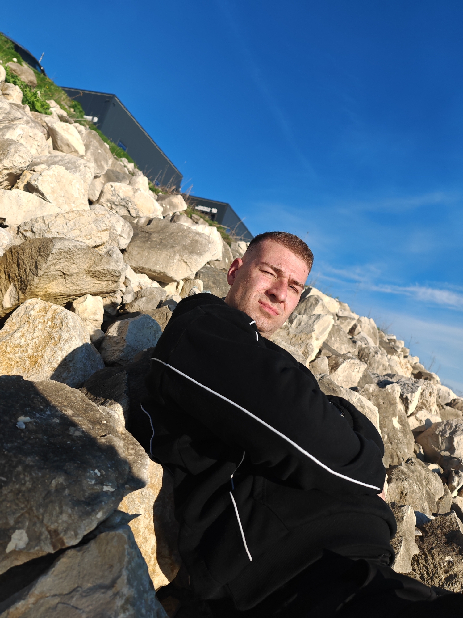 The image captures a man with a buzzcut, dressed in a black tracksuit with white stripes, leaning against a pile of large, light-colored rocks. The setting appears to be outdoors under a bright blue sky, possibly near a hillside with a building visible in the background. The location is Shumen, Bulgaria.