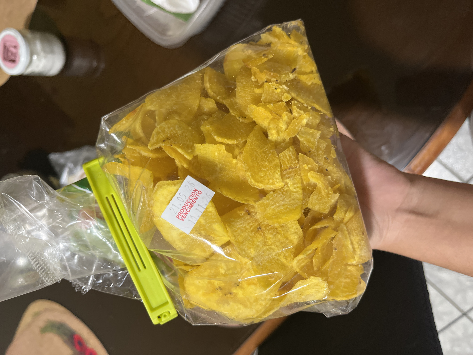A close-up, eye-level shot features a hand holding a transparent plastic bag full of bright yellow, thinly sliced chips, likely plantain or banana chips. The chips vary in shape and size, displaying some darker specks, possibly seasoning or slight browning from frying. The bag is sealed at the top with a prominent neon green plastic clip.
Affixed to the front of the bag is a small white rectangular sticker with red text. The visible text reads: "21.05.25", followed by "PRODUCCION", and then "VENCIMIENTO 10.11.25", indicating production and expiration dates.
The background is mostly out of focus, revealing a dark, reflective surface that appears to be a table. To the left, a white bottle with a pink lid is partially visible, along with a clear plastic container and another plastic bag containing green, leafy contents, also secured with a similar green clip. On the right, a glimpse of a person's leg and part of a tiled floor suggests an indoor setting, such as a kitchen or dining area. The scene is illuminated by ambient indoor lighting. Given the context of San Luis, Peru, these chips are a typical local snack.