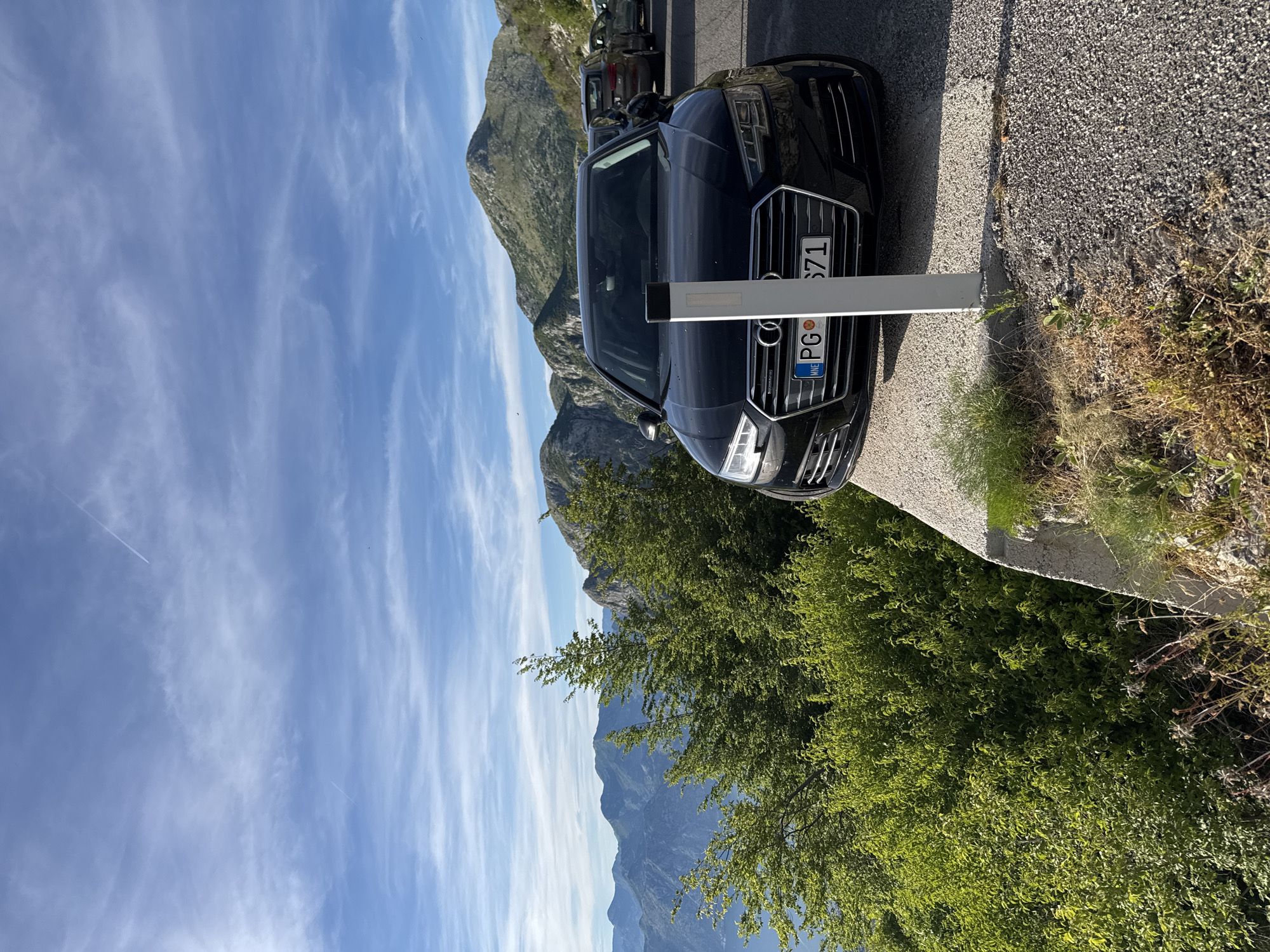 A black Audi A4 sedan is parked on the side of a winding mountain road with a panoramic view of mountains and a body of water. The car is in the foreground, facing towards the viewer, with its front bumper and license plate clearly visible. The license plate reads "PG 571". The road appears to be paved with asphalt and gravel. To the left of the car, lush green trees and foliage cascade down the mountainside, obscuring the lower half of the view. In the background, a range of rugged mountains stretches across the horizon, with a clear blue sky dotted with wispy clouds above. The scene suggests a scenic overlook in the mountainous region of Žanjev Do, Montenegro, likely a popular stopping point for travelers.