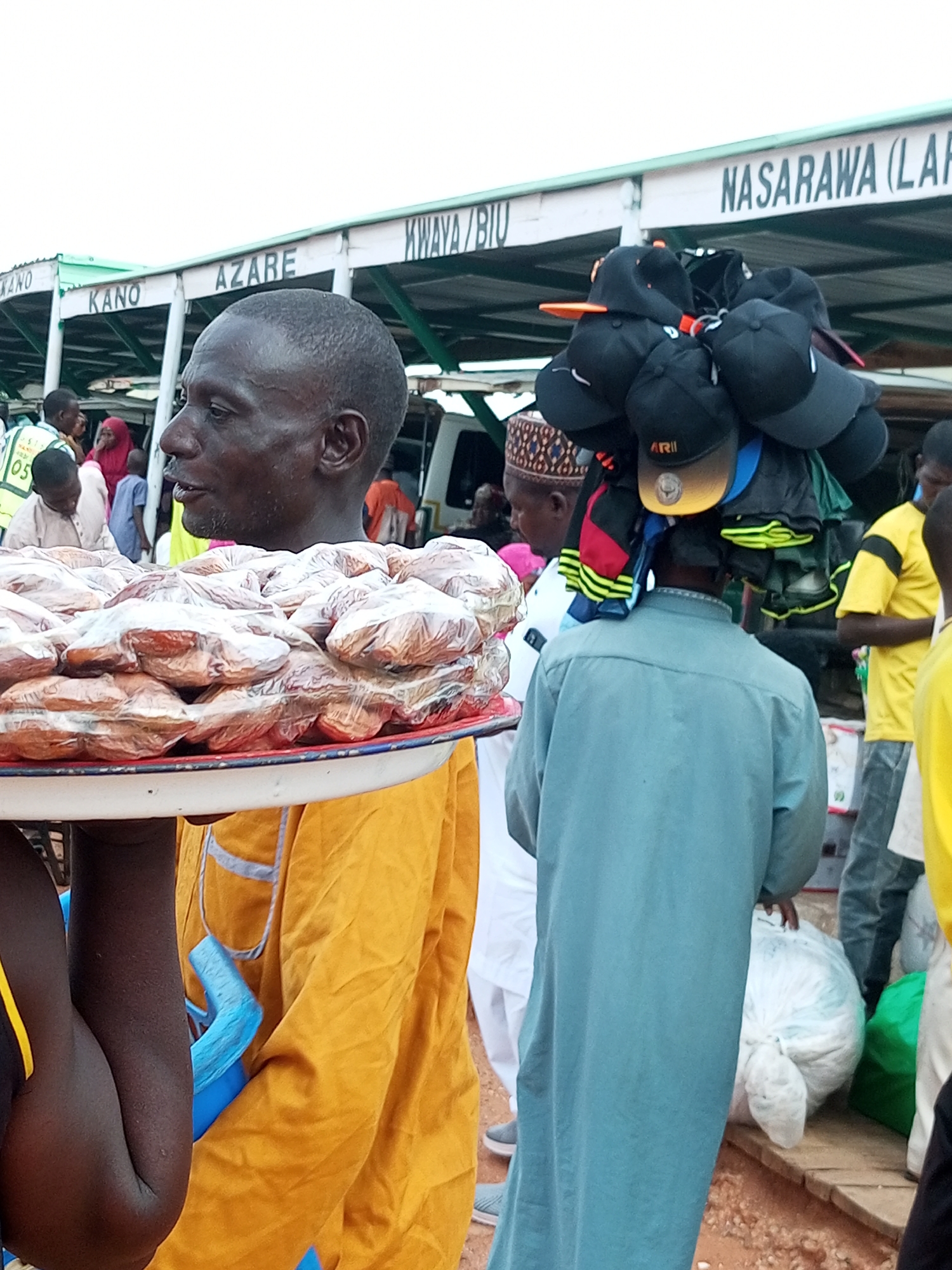 In Gombe, Nigeria, a street vendor in a yellow robe presents a tray of individually plastic-wrapped baked goods. Behind him, another vendor carries a selection of baseball caps on his head, his back to the camera, dressed in a blue robe. The scene unfolds near a marketplace stall with signage indicating various locations, including "KANO," "AZARE," and "KWANYA/BIU," and "NASARAWA (LAR." A white minivan is partially visible. Several other individuals are present in the background, some wearing traditional attire. The ground is a mixture of red earth and wooden planks. The lighting suggests an overcast day.
