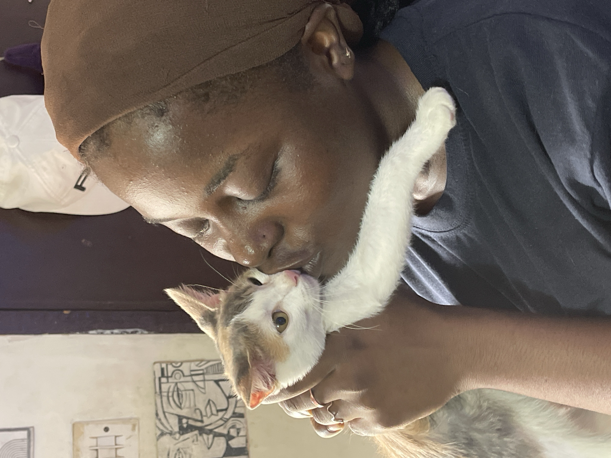This media file features a close-up shot of a person with dark skin, wearing a brown headscarf and a dark t-shirt, gently holding and kissing a calico kitten. The kitten has white, brown, and gray patches, with large amber eyes and its tongue extended, seemingly licking the person's nose. The person's eyes are closed, and they appear to be experiencing a moment of affection and tenderness.
In the background, a white baseball cap is visible, and the overall setting suggests an indoor environment. The lighting is soft, indicating it could be daytime, though no specific time is discernible. The image is framed to focus on the intimate interaction between the person and the kitten, highlighting the bond between them. There is no visible text in the image. The scene is one of warmth and care.