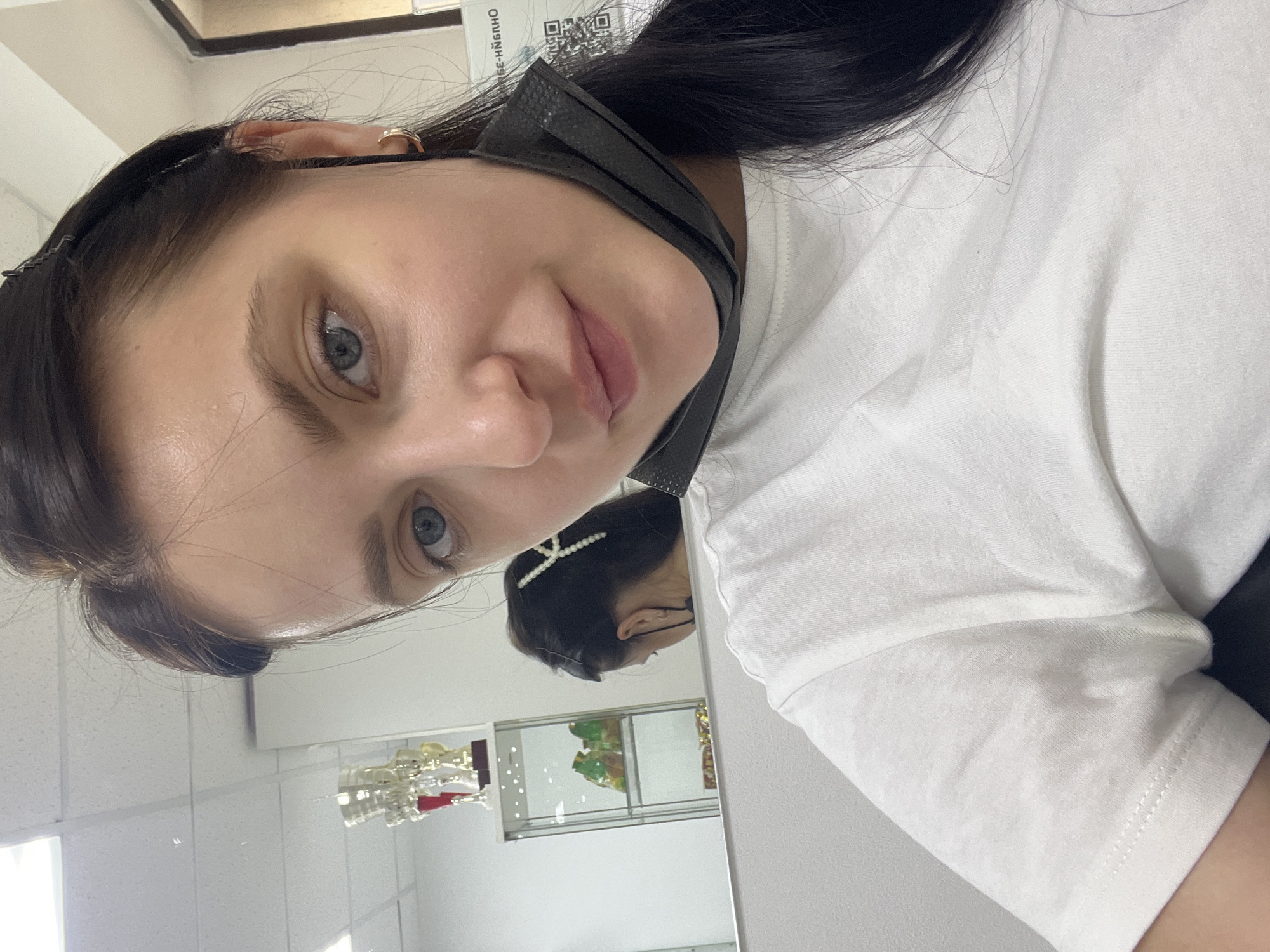 A woman with dark hair and blue eyes is captured in a close-up, upward-angled shot. She is wearing a white short-sleeved t-shirt and has a black face mask pulled down under her chin. Her dark hair is styled with small clips near her forehead and a decorative pearl hair clip visible on the back of her head in the mirror's reflection. A small, golden-colored ring is visible on her left earlobe. She looks directly at the camera with a neutral to slightly pleasant expression.
The background shows a brightly lit indoor setting, likely a commercial space in Chaikovsky, Russia. A large mirror reflects the back of the woman's head and a section of the room behind her. In the mirror's reflection, display cases containing various items, possibly jewelry or decorative figurines, are visible. Above the woman's head, in the background, a white sign or paper with Cyrillic text and a QR code is partially visible. The text appears to read "ОНЧЙБНЭ-39". The overall lighting suggests it is daytime.
