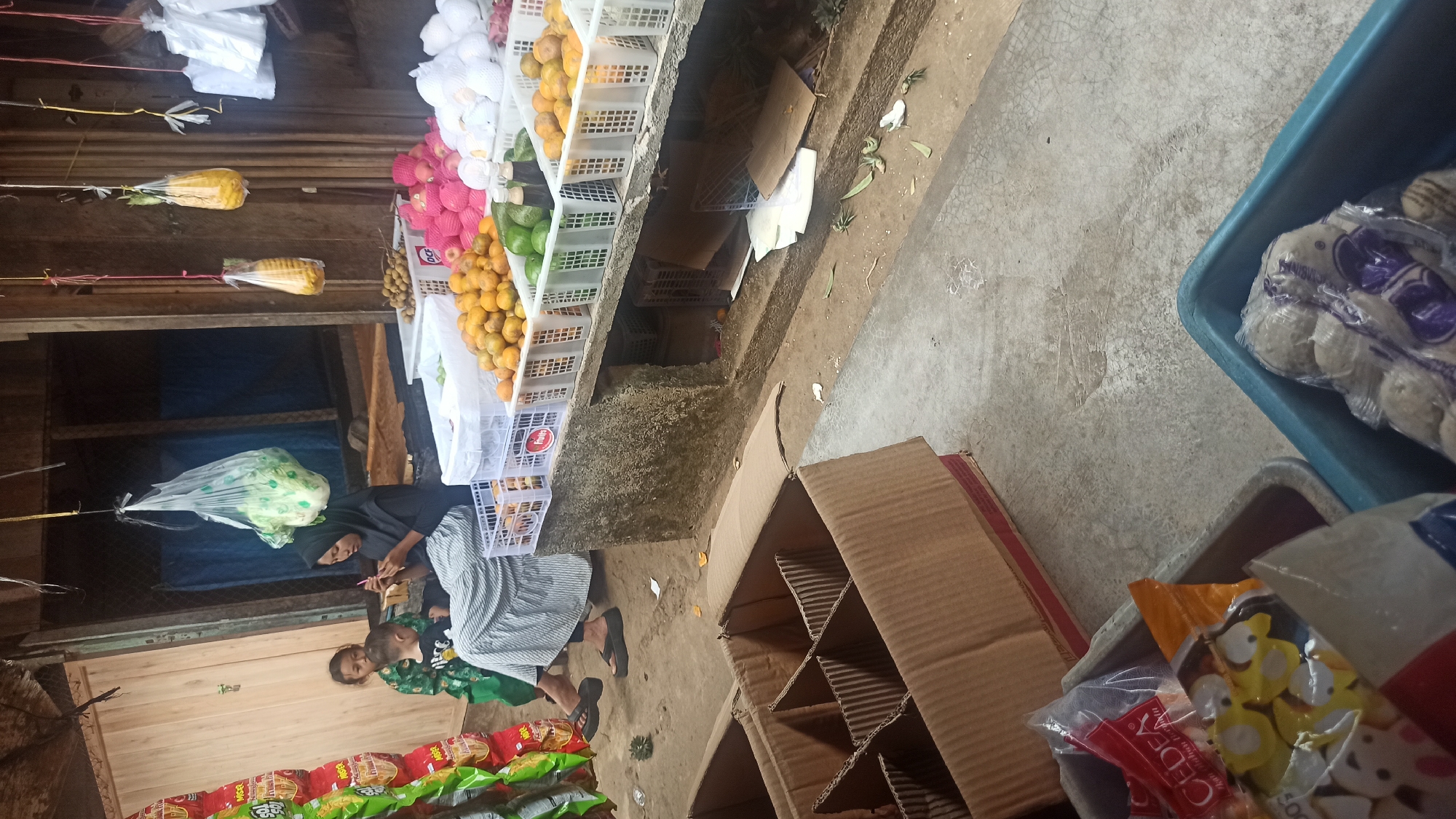 The image depicts an outdoor market scene in Tuba, Indonesia. In the background, a woman wearing a hijab and a long skirt is seated, holding a young child. Beside them, another child stands. The woman appears to be engaged with her phone or a similar device. In front of them, a counter displays an assortment of fruits in white plastic crates, including oranges, longans, apples, and avocados. Hanging from the ceiling are bags of produce, including what appears to be lettuce and what might be dried corn or some other grain. The foreground shows several cardboard boxes, one of which is open and divided into compartments. A plastic tub containing bags of meatballs is also visible in the lower right corner, alongside another tub with packages of snacks. The ground is concrete, and there are some scattered leaves and debris. The overall setting suggests a local, informal marketplace.