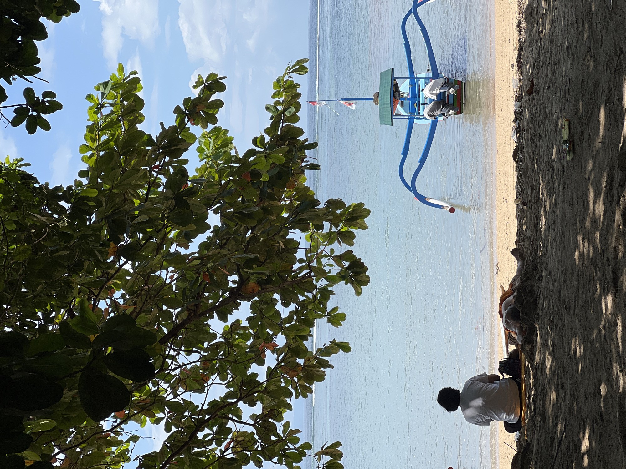 This image, taken in Denpasar, Indonesia, captures a sunny tropical beach scene from beneath the shade of a large tree. The photo is oriented vertically, showing the ocean to the right and sandy beach to the left, with lush green foliage from a tree framing the top and bottom.
In the mid-ground, a traditional Indonesian outrigger boat, known as a jukung, with a blue hull and outriggers, floats in the calm, clear turquoise water near the shore. Two people appear to be seated within the boat, which also features a small canopy and a red and white flag, possibly resembling the Indonesian flag, on a pole.
On the sandy beach in the foreground, a person with dark hair, wearing a white t-shirt, sits facing the water, their back to the viewer. A bag or backpack is visible beside them. Further along the shaded beach, another person is lying down, partially obscured by shadows. The sandy area shows some scattered debris, including a plastic bottle and cup.
The time of day appears to be bright daylight, possibly morning or early afternoon, given the clear blue sky with scattered white clouds visible through the tree canopy, and the strong shadows cast by the tree on the sand. The atmosphere is one of peaceful relaxation on a scenic beach.