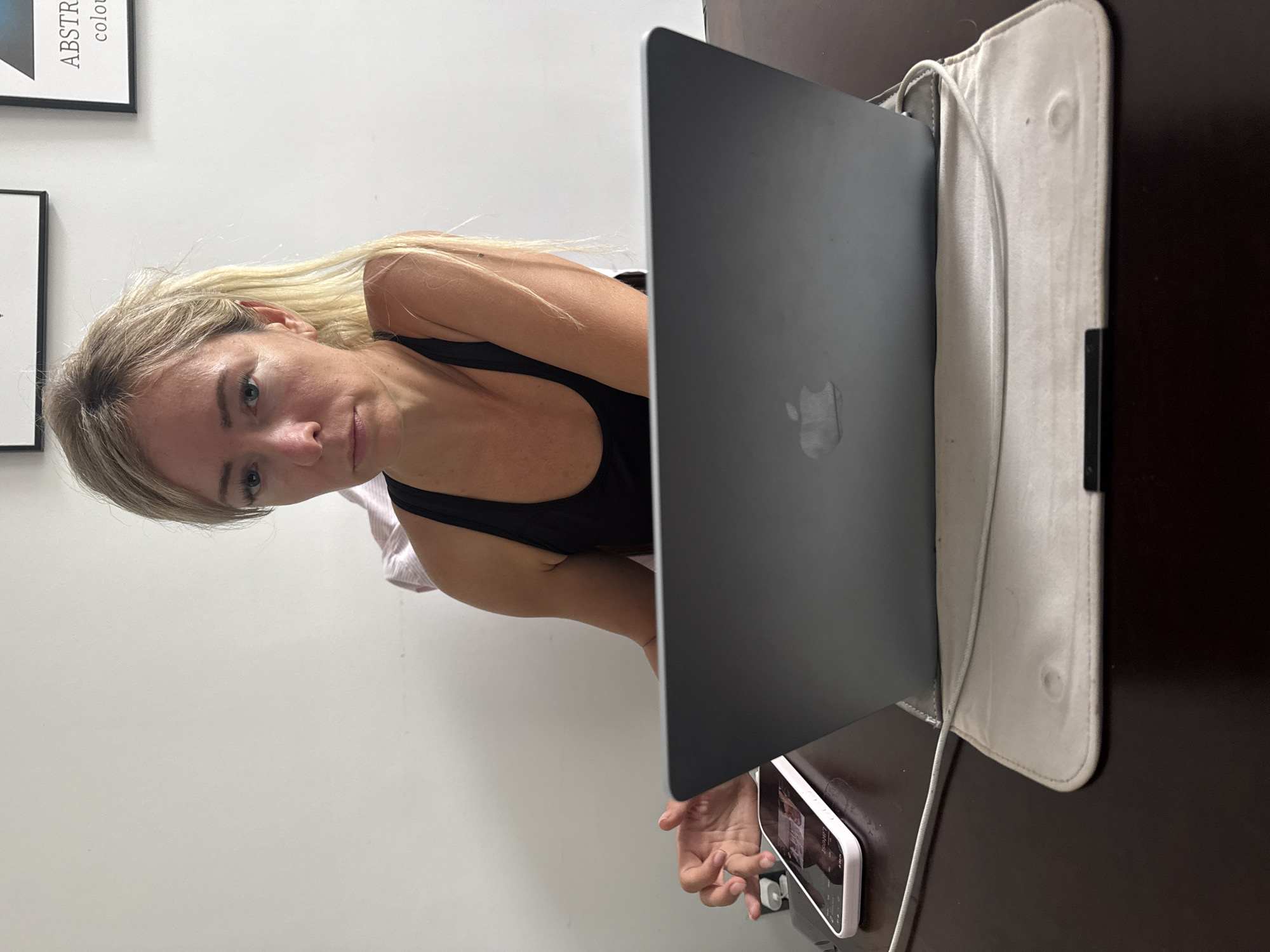 An overhead shot shows a woman with blonde hair in a ponytail sitting at a dark wooden desk. She is wearing a black tank top and looking directly at the camera with a neutral expression. In front of her is a silver MacBook laptop, open and resting on a light-colored protective sleeve. To the left of the laptop is a white smartphone with its screen displaying what appears to be a video. The woman's left hand is extended, with her index finger touching the phone screen. A white power cord runs from the laptop, over the sleeve, and towards the left side of the frame. The background consists of a plain white wall. There are two framed pictures on the wall behind the woman. This scene is set indoors, at a desk in what appears to be a home or office environment, possibly in Xuân Trường - Đà Lạt, Vietnam, given the context.