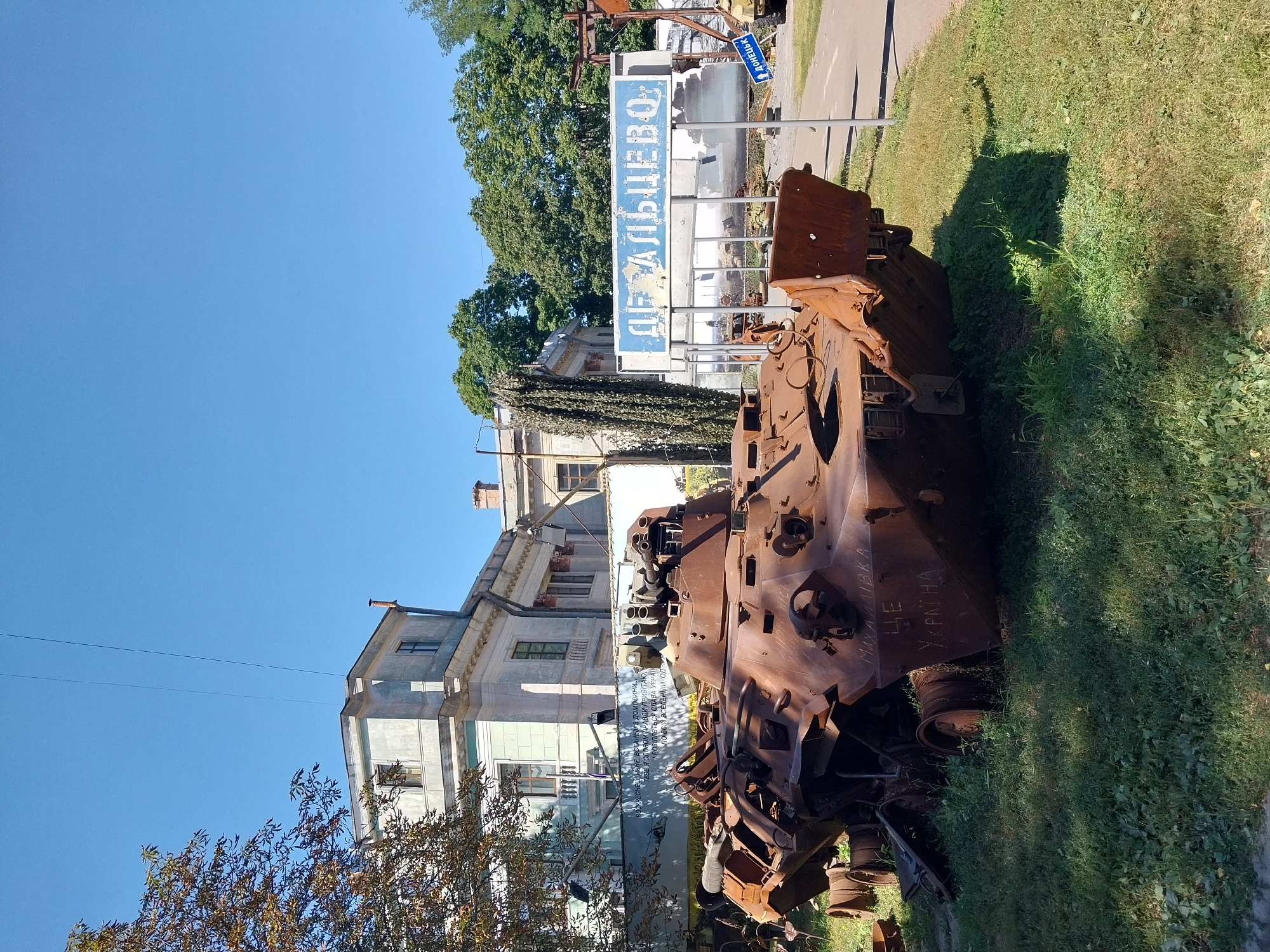 The image depicts a memorial site in the city of Dnipro, Ukraine, dedicated to victims of the conflict. In the foreground, a heavily damaged and rusted armored personnel carrier (APC) rests on grassy ground. The APC shows significant signs of combat damage, with sections of its hull and turret appearing blown open or melted. White Cyrillic lettering is visible on the side of the APC, reading "МАХАЧКАЛА" and below it "ЦЕ УКРАЇНА" (This is Ukraine).
Behind the APC, a light blue sign with white Cyrillic lettering reads "ДЕБАЛЬЦЕВО". This sign refers to the Battle of Debaltseve, a significant engagement during the war in Donbas. Further back, a white building with classical architectural elements stands, likely a museum or historical structure. A banner with text in Ukrainian and imagery related to the conflict is also visible behind the APC. The environment is outdoors under a clear blue sky, with trees in the background. The overall scene conveys a somber atmosphere, serving as a historical marker of the war.