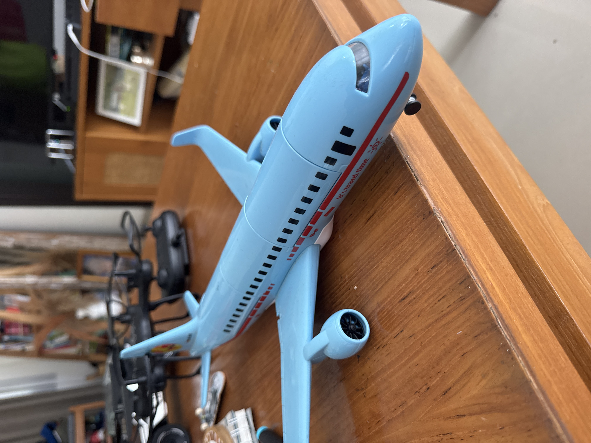 A light blue toy airplane rests diagonally on a wooden surface in an indoor setting. The airplane, which appears to be a model or child's toy, features black window decals and a prominent red stripe along its fuselage. White text within the red stripe reads "It's travel time," accompanied by a small sun graphic. The airplane has two engines mounted under its wings.
The wooden surface beneath the plane exhibits a distinct grain and a warm, light brown color, suggesting it is a table or shelf. In the blurred background to the left, dark objects resembling electronic equipment or furniture are visible, alongside wooden shelving units containing various unidentifiable items. One small, framed picture is discernible on a shelf. The lighting indicates an interior environment. No specific activity is depicted, only the static display of the airplane.