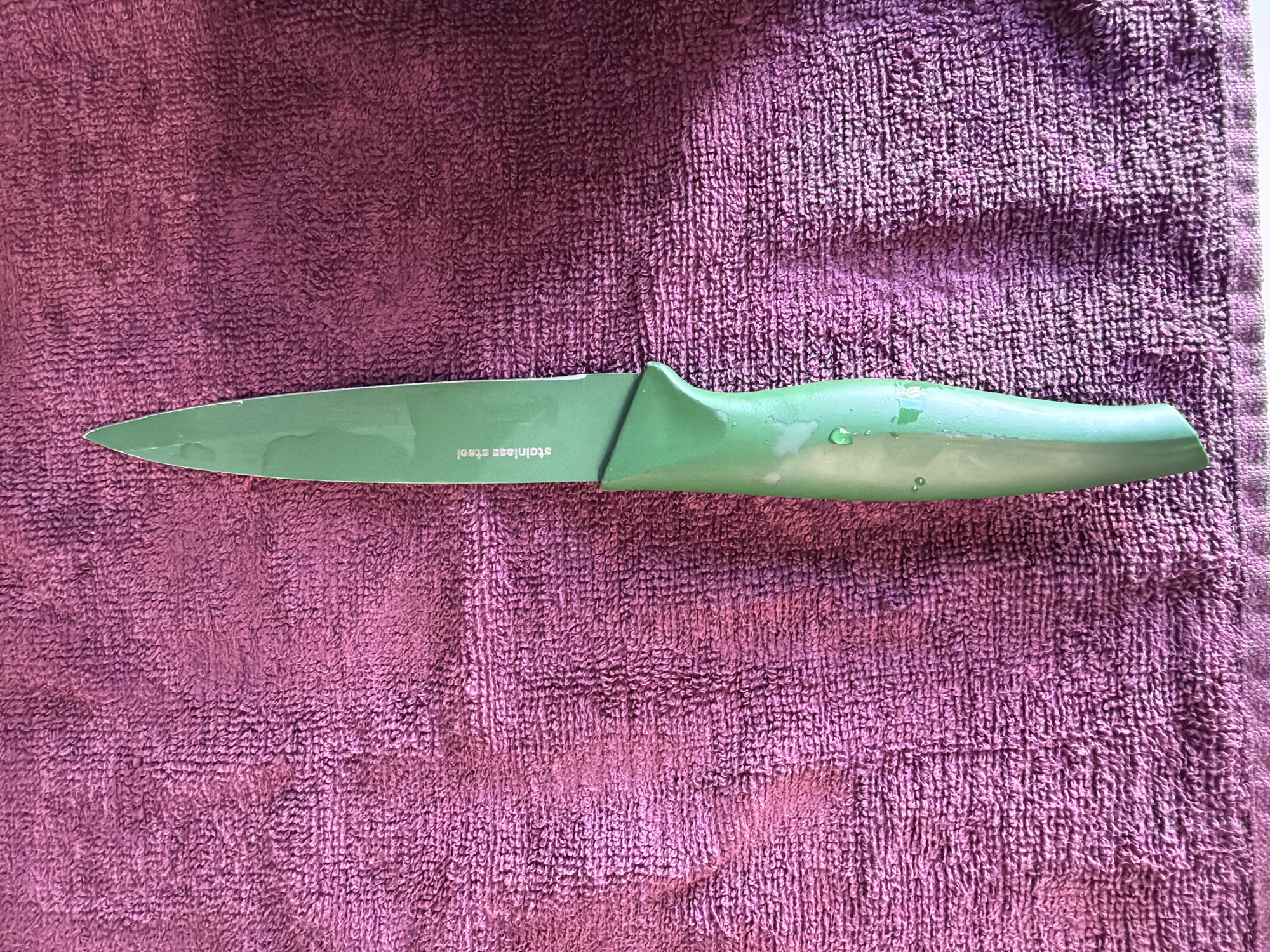 The image shows a green paring knife lying on a purple towel. The knife is oriented horizontally in the center of the image. The blade is imprinted with the phrase "stainless steel". There is no evidence to suggest that this image was taken in Arinj, Armenia. No people, subjects, objects, actions, or interactions can be discerned other than the knife resting on the towel. The environment is limited to the towel and the knife.