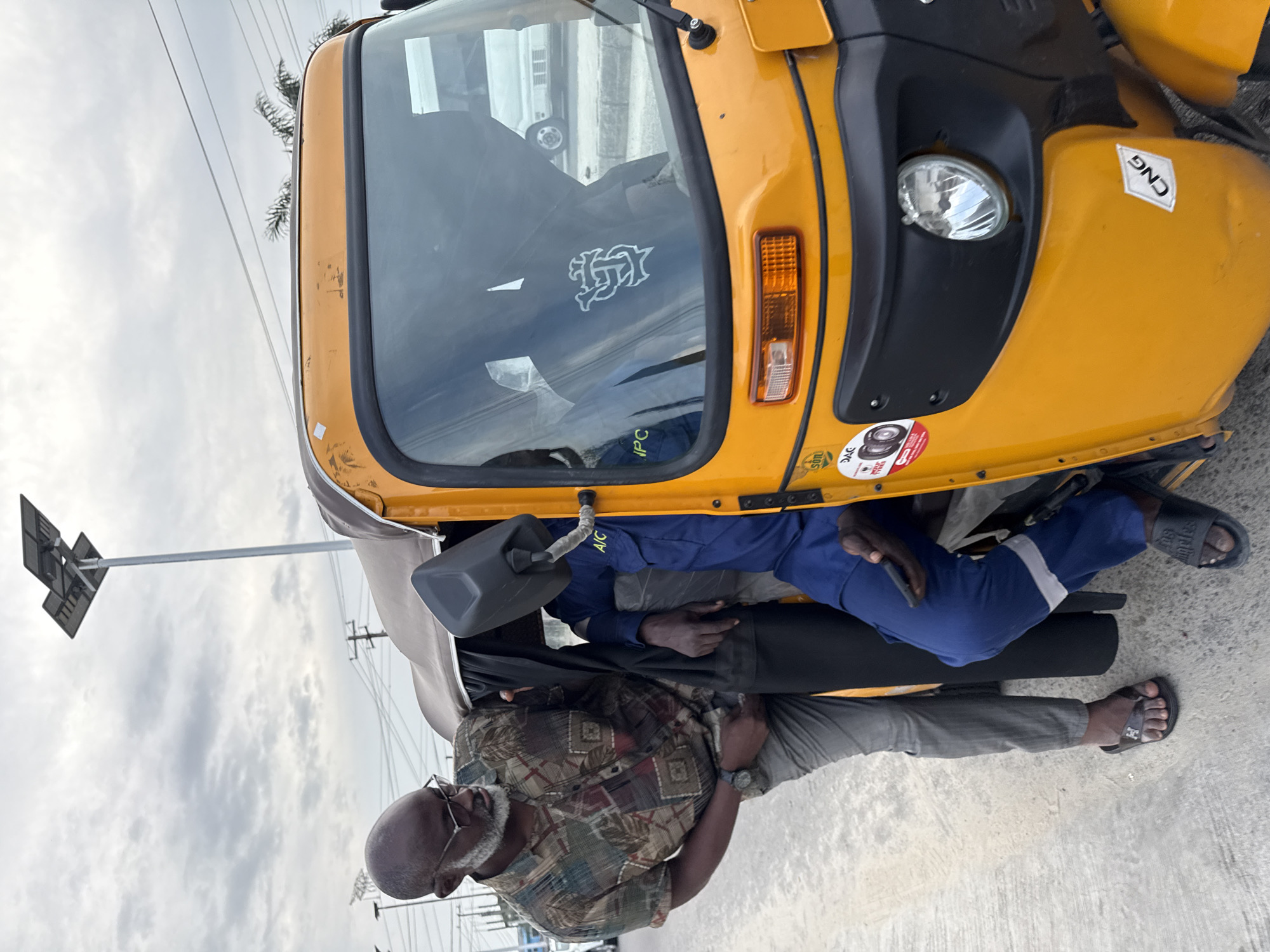 In Ado, Nigeria, a man in a patterned shirt and grey pants, wearing glasses and a beard, is leaning into a yellow auto-rickshaw. He appears to be interacting with another person inside the vehicle who is wearing a blue uniform with a white stripe and the letters "A/C" visible. The auto-rickshaw has a dark brown canopy and a black side mirror. The scene takes place on a paved surface, possibly a street, under an overcast sky. In the background, there are power lines and streetlights, including one with solar panels. The auto-rickshaw displays several stickers, including one with "CNG" on the front fender.