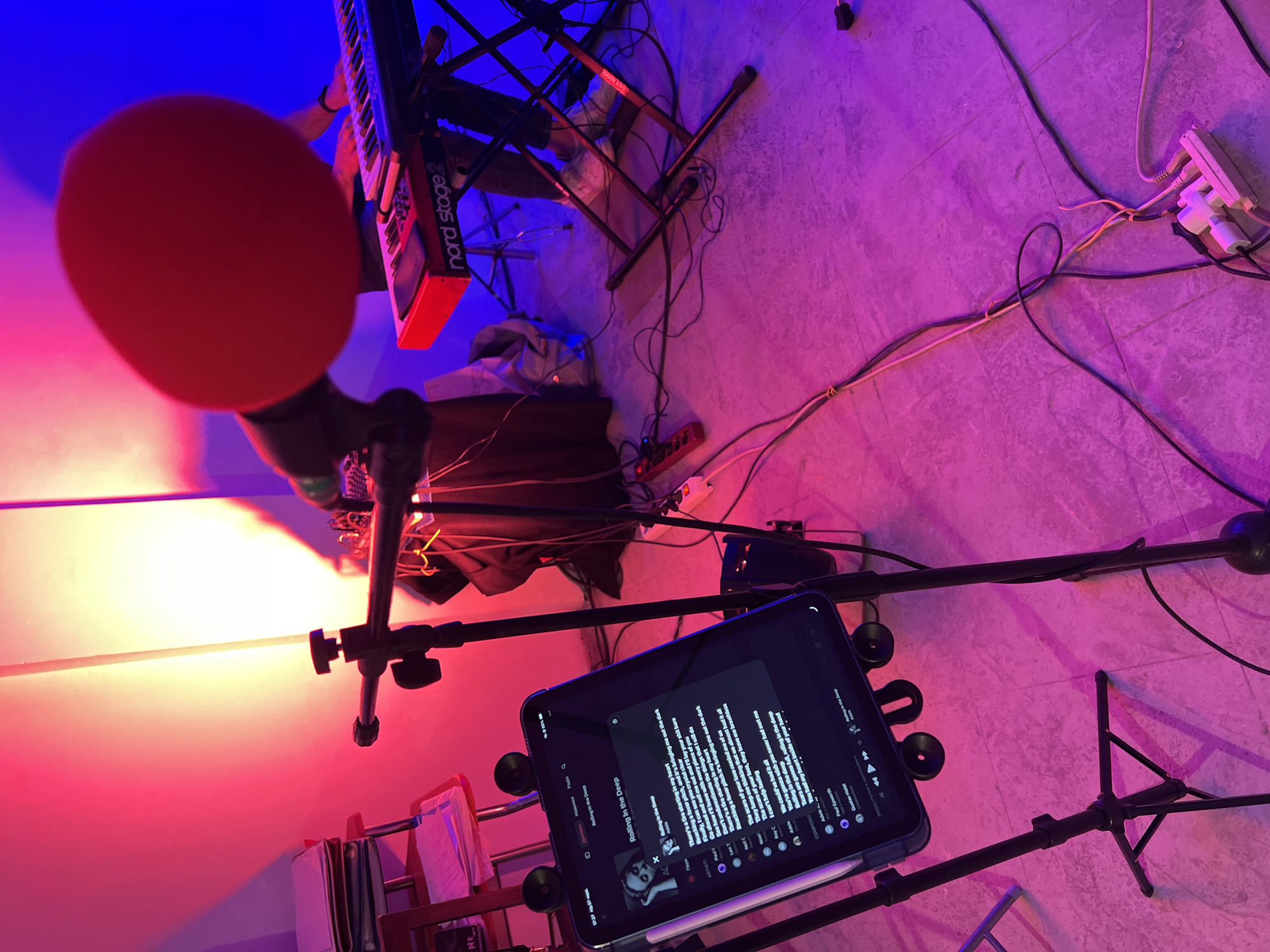 The image depicts an indoor space, identified as being in Santeramo in Colle, Italy, configured for a musical activity. The room is illuminated by prominent red and blue ambient lighting, casting magenta and purple hues on the tiled floor.
In the background, a single individual is partially visible. Their upper body and head are obscured by a large, spherical red object, likely a microphone pop filter or windscreen, positioned centrally. The individual's hands are visible on the keys of a red Nord Stage 2 keyboard, which rests on a black X-style stand. Their lower legs and feet, wearing white sneakers, are visible beneath the keyboard stand.
To the left foreground, a black tablet, encased and mounted on a microphone stand, displays lyrics for Adele's "Rolling In The Deep," along with artist and composer information. A stylus is attached to the tablet's side. Adjacent to the tablet stand, a small, light-colored chair with a reddish frame holds a stack of papers or documents.
The floor is a light-colored tile, with numerous black, white, and yellow cables distributed across its surface; some are bundled with ties. Two power strips are visible on the floor: a red one positioned on the left side near the keyboard stand, and a white one on the far right, both with multiple plugs inserted.
The background wall exhibits distinct color zones, with red light illuminating the left side and blue light illuminating the right side behind the keyboard setup. The overall environment suggests a functional music practice or performance space with multiple electronic devices connected.
