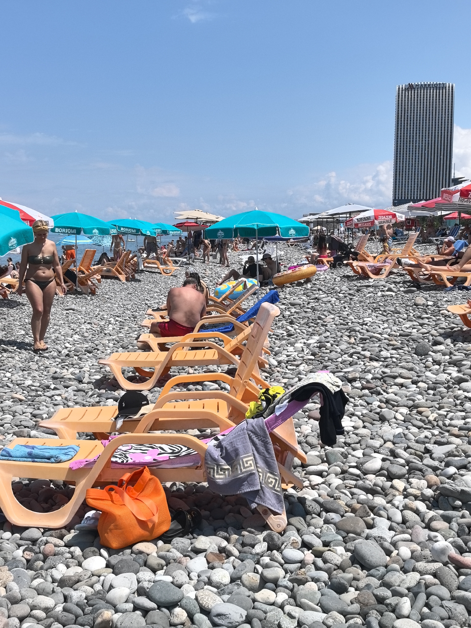 A sunny day at a pebble beach in Batumi, Georgia, shows numerous beachgoers enjoying the weather. In the foreground, several empty orange plastic lounge chairs are arranged on the rocky shore, with towels and bags placed on them. A woman in a bikini walks away from the camera towards the right, while a shirtless man in red shorts sits on a lounge chair in the middle ground. In the background, more people are scattered across the beach, some under umbrellas, and a tall, modern skyscraper dominates the skyline. The sea is not visible in this frame.